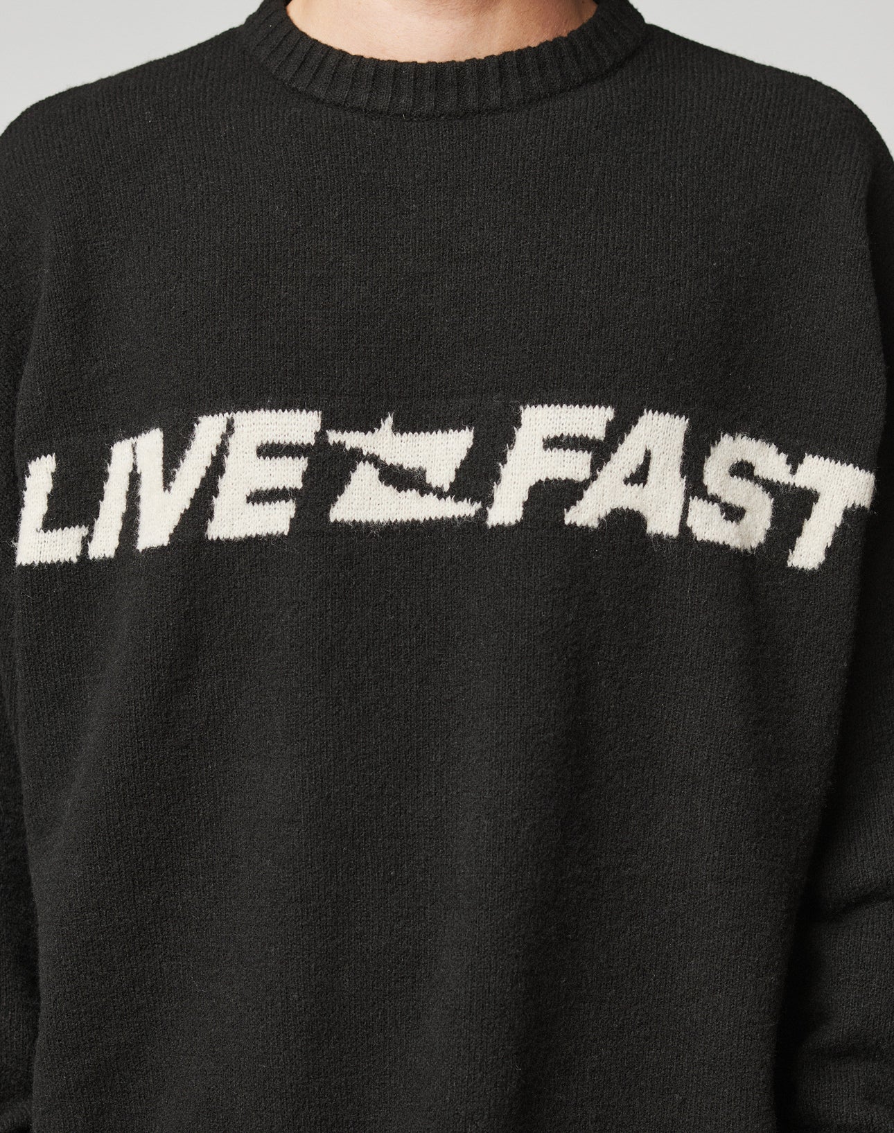 A person wears the Live Fast Boucle Sweater by LIVE FAST DIE YOUNG—an oversized black piece with LIVE FAST and a fast-forward arrow in bold white, crafted from soft Strick polyester for comfort and style.