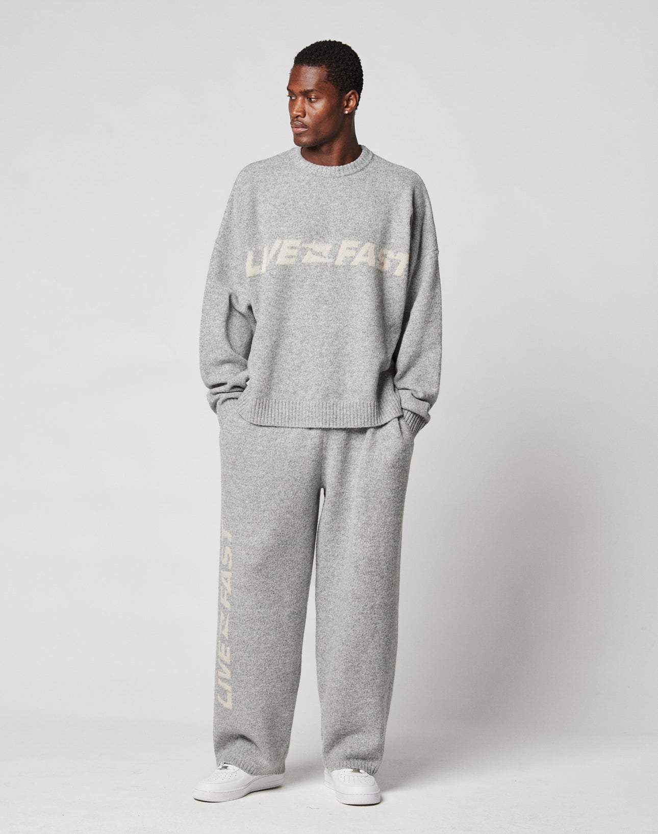 A man stands against a plain background in an oversize Live Fast Boucle Sweater and matching sweatpants from LIVE FAST DIE YOUNG, both light gray with “LIVE FAST” printed in white. He completes the look with white sneakers.