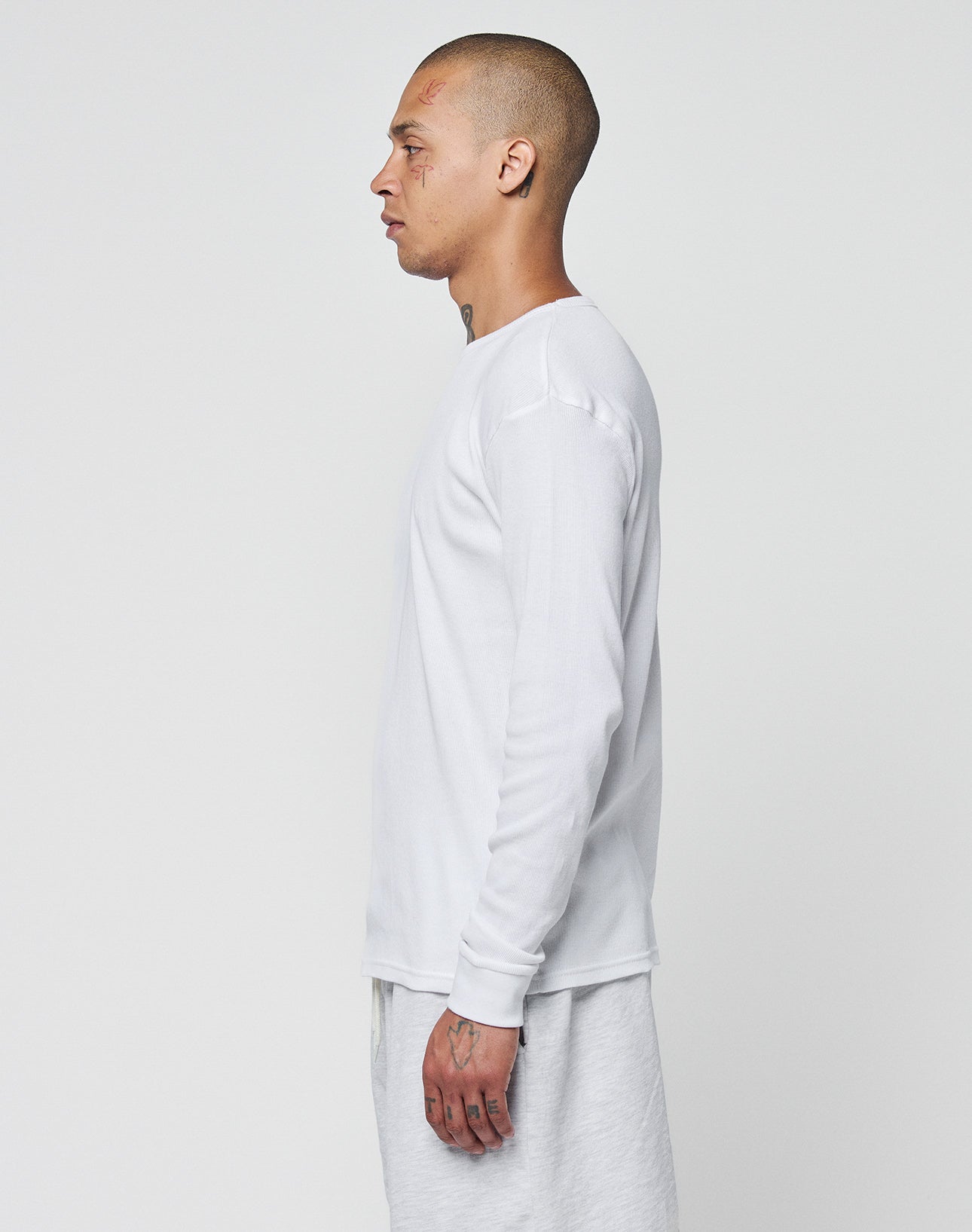 A person with a shaved head and visible tattoos stands in profile against a white background, wearing the LIVE FAST DIE YOUNG Basic Regular Rib Longsleeve in Regular Fit and light-colored pants, both made from soft Baumwolle for comfort.
