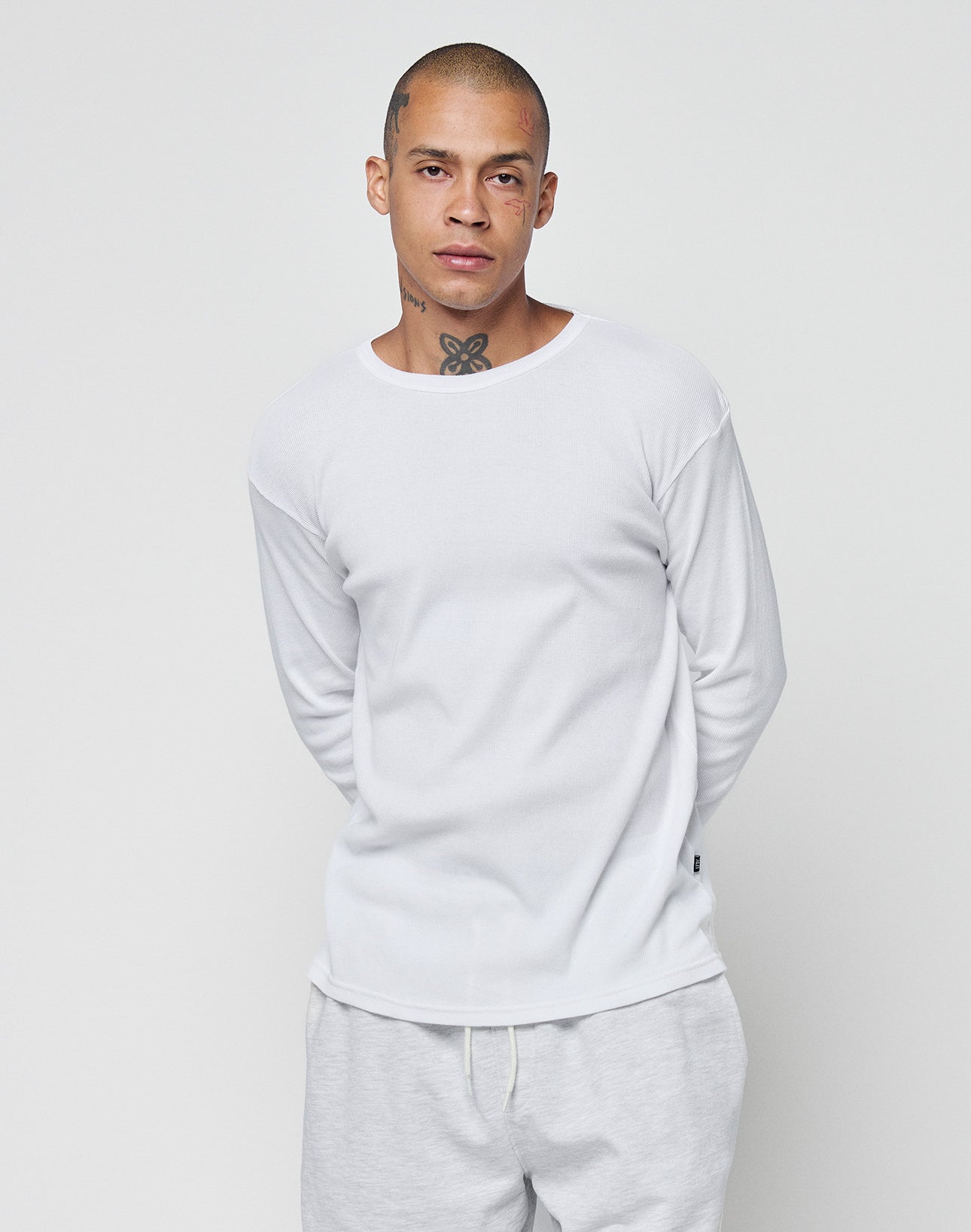 A person with a shaved head and visible neck and face tattoos wears the LIVE FAST DIE YOUNG Basic Regular Rib Longsleeve in white, paired with light-colored pants, standing against a plain white background with hands behind their back.