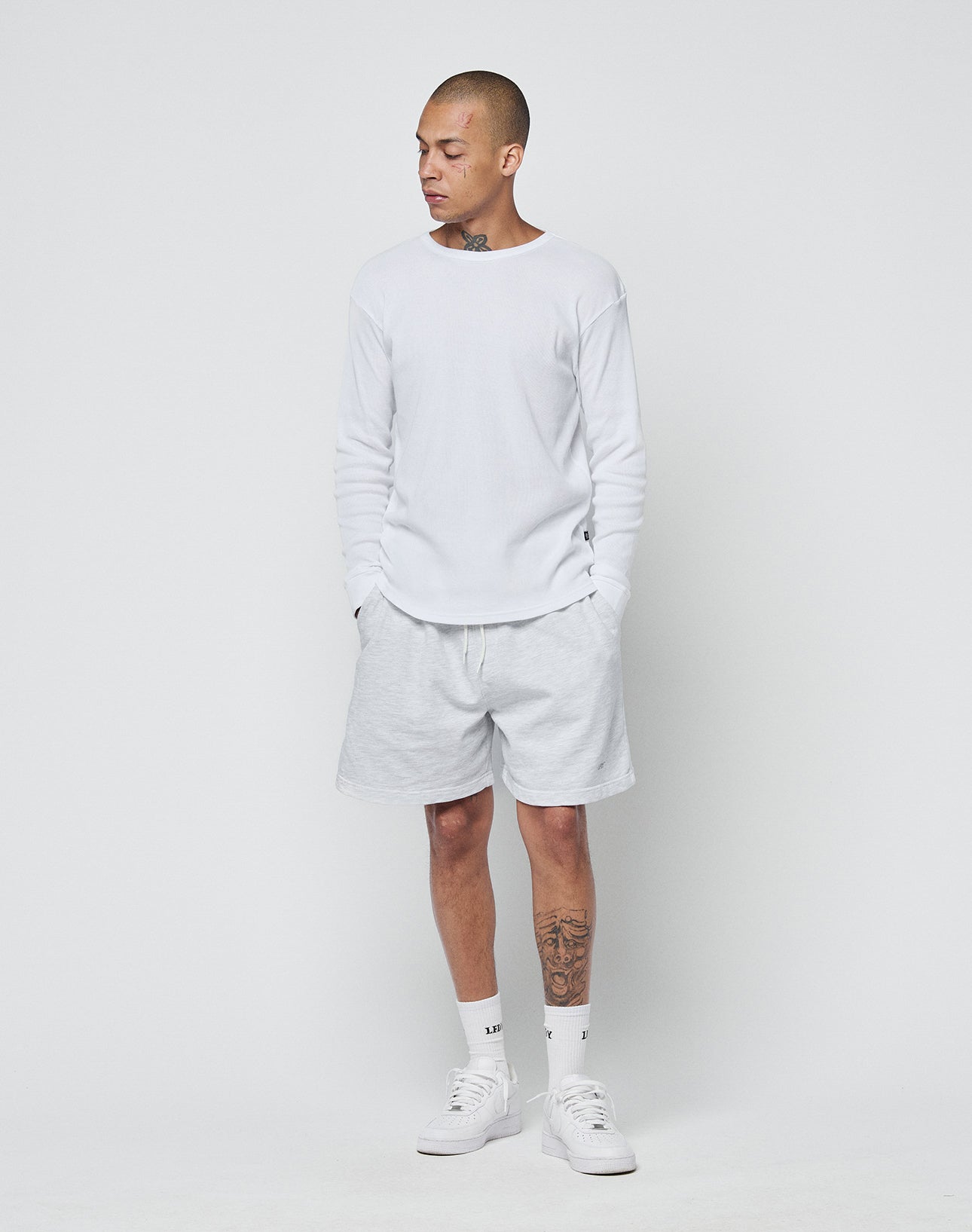 A person stands against a plain white background wearing a LIVE FAST DIE YOUNG Basic Regular Rib Longsleeve in white, white shorts, socks, and sneakers with hands in their shorts pockets. Tattoos are visible on their neck and left leg.