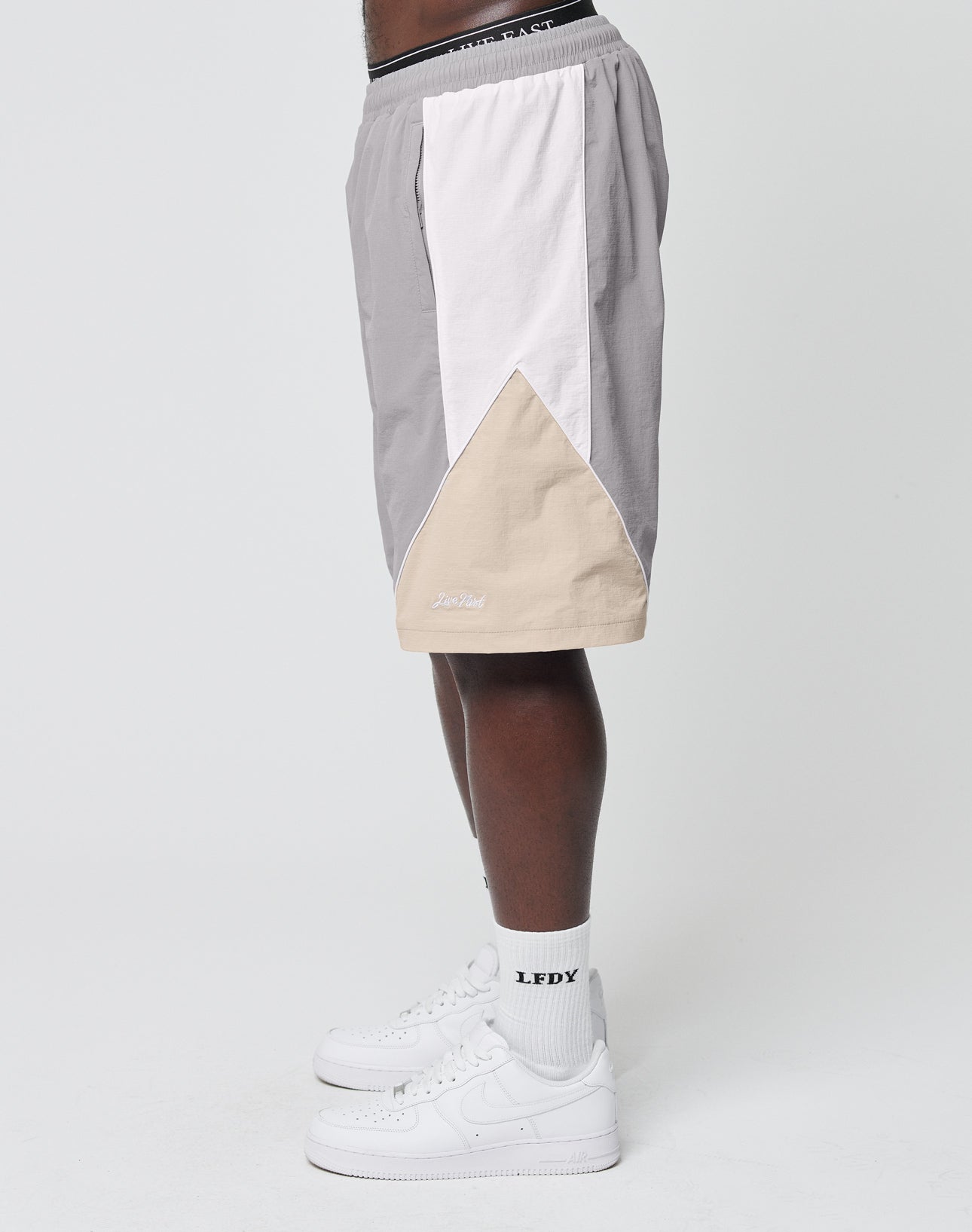 A person wears LIVE FAST DIE YOUNG Basic Nylon Trackshorts in color-block gray, white, and beige with an elastic waistband and regular fit, paired with white LFDY socks and white sneakers, standing sideways against a plain white background.