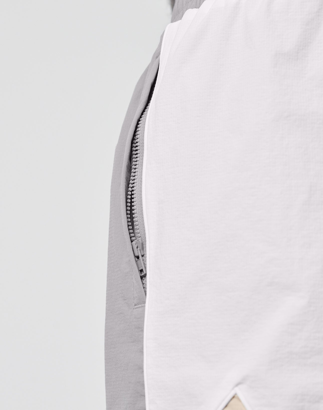 Close-up of LIVE FAST DIE YOUNG Basic Nylon Trackshorts in Regular Fit, showing a partially visible zipper between light gray and white Nylon-Lycra fabric, with a small beige triangle detail at the bottom edge.