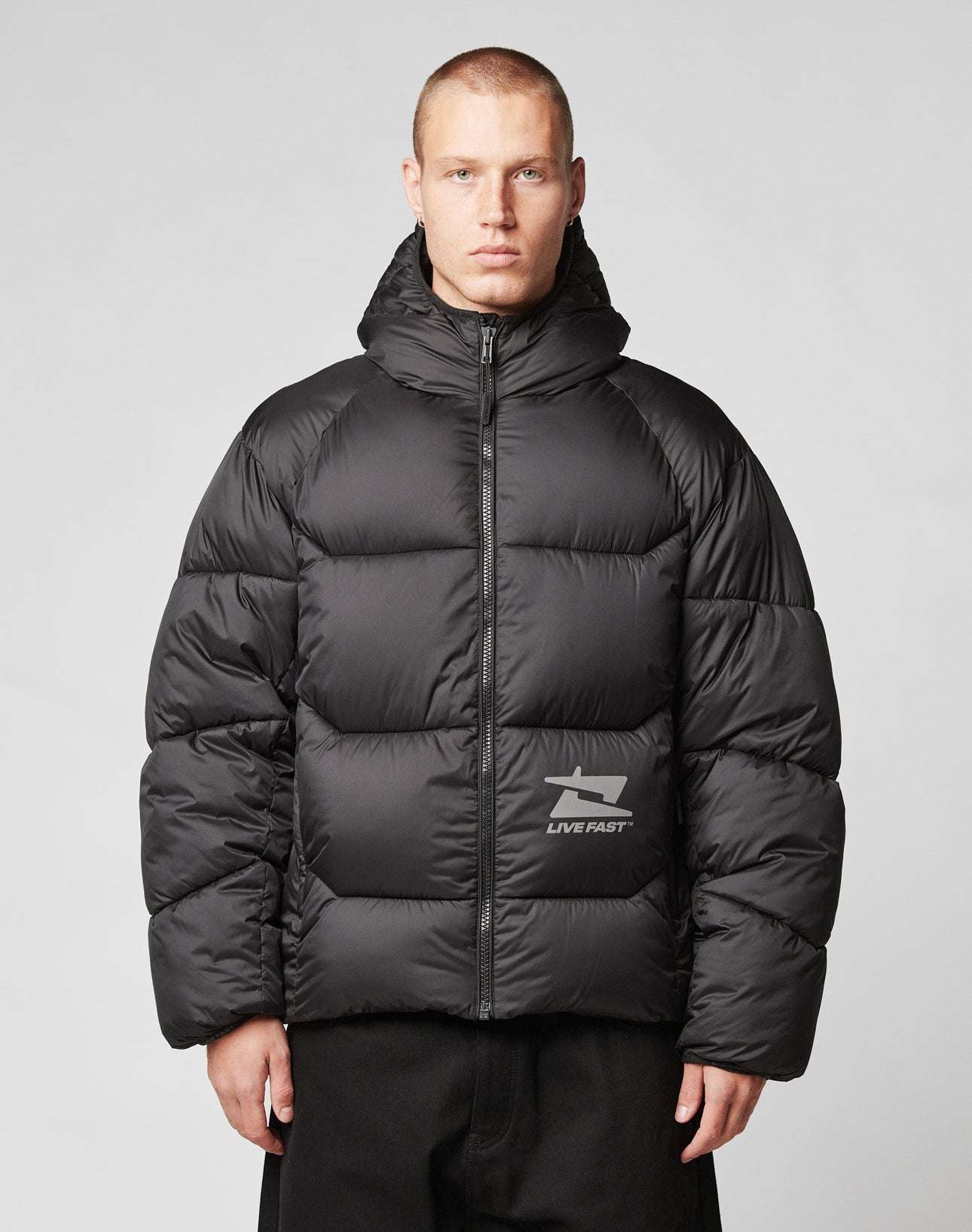 A person with short hair wears the LIVE FAST DIE YOUNG Live Fast Puffer Jacket—black, hooded, water-resistant, and featuring a reflective LIVE FAST print on the left chest—against a plain light gray background.