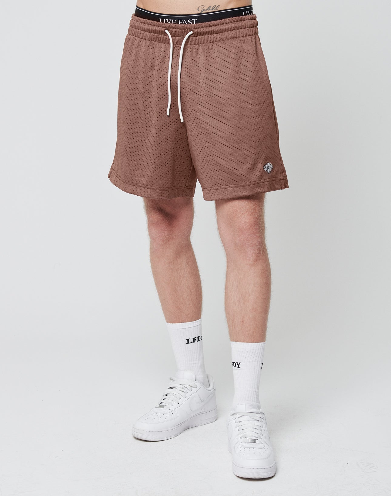 A person stands against a plain white background wearing LIVE FAST DIE YOUNG Basic Mesh Shorts in light brown, white socks with black text, white sneakers, and a black waistband with white lettering and a logo patch. The upper body is not visible.