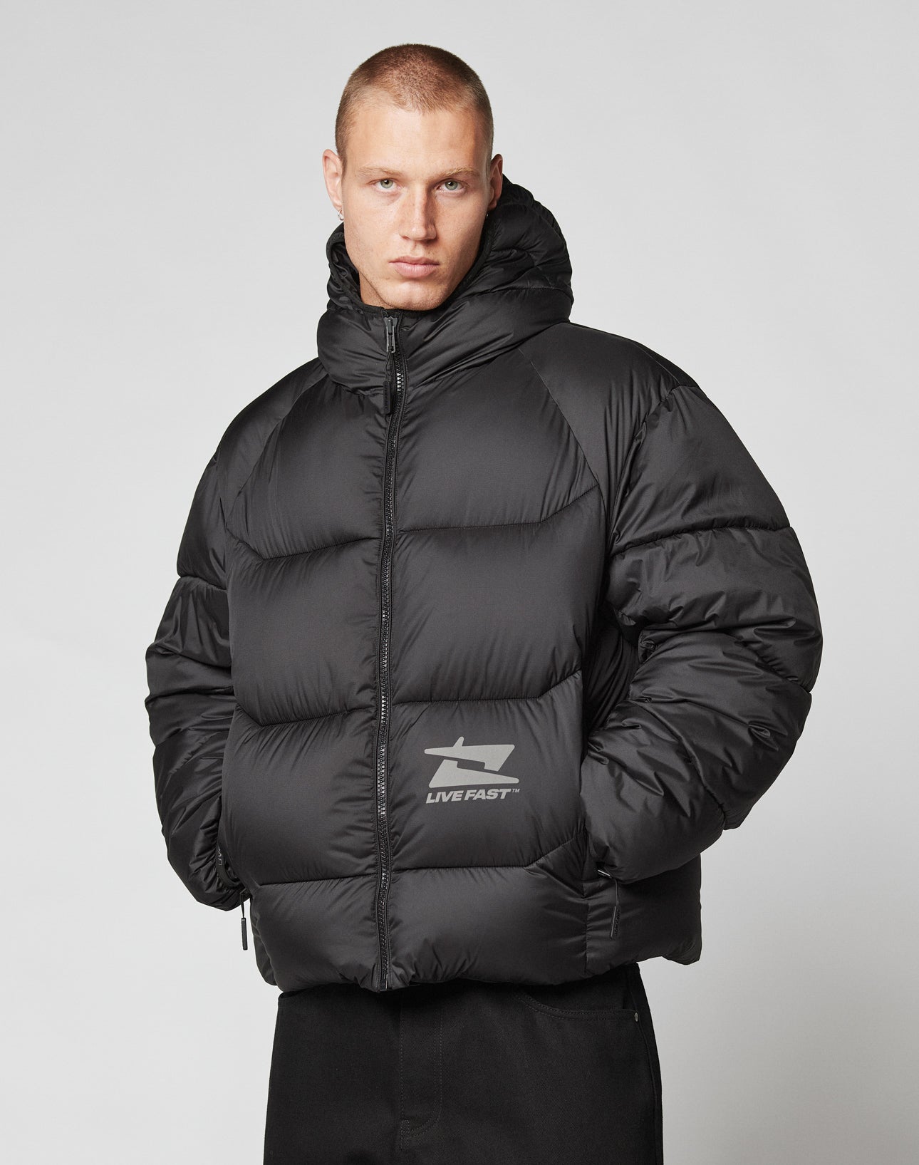 A person in the oversize fit Live Fast Puffer Jacket by LIVE FAST DIE YOUNG, featuring a hood and reflective LIVE FAST logo on the front, stands against a plain light gray background with hands in the water-repellent jackets pockets.