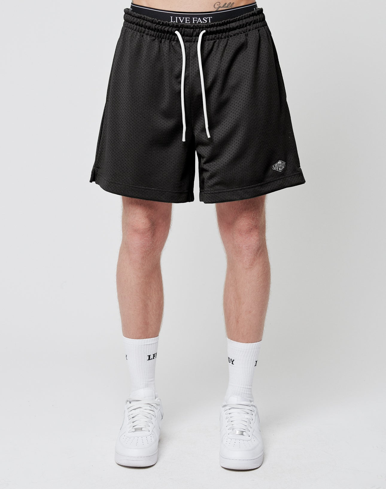 A person models the LIVE FAST DIE YOUNG Basic Mesh Shorts—black polyester with white drawstrings—paired with white socks and sneakers, against a plain white background. Only their lower half is shown.