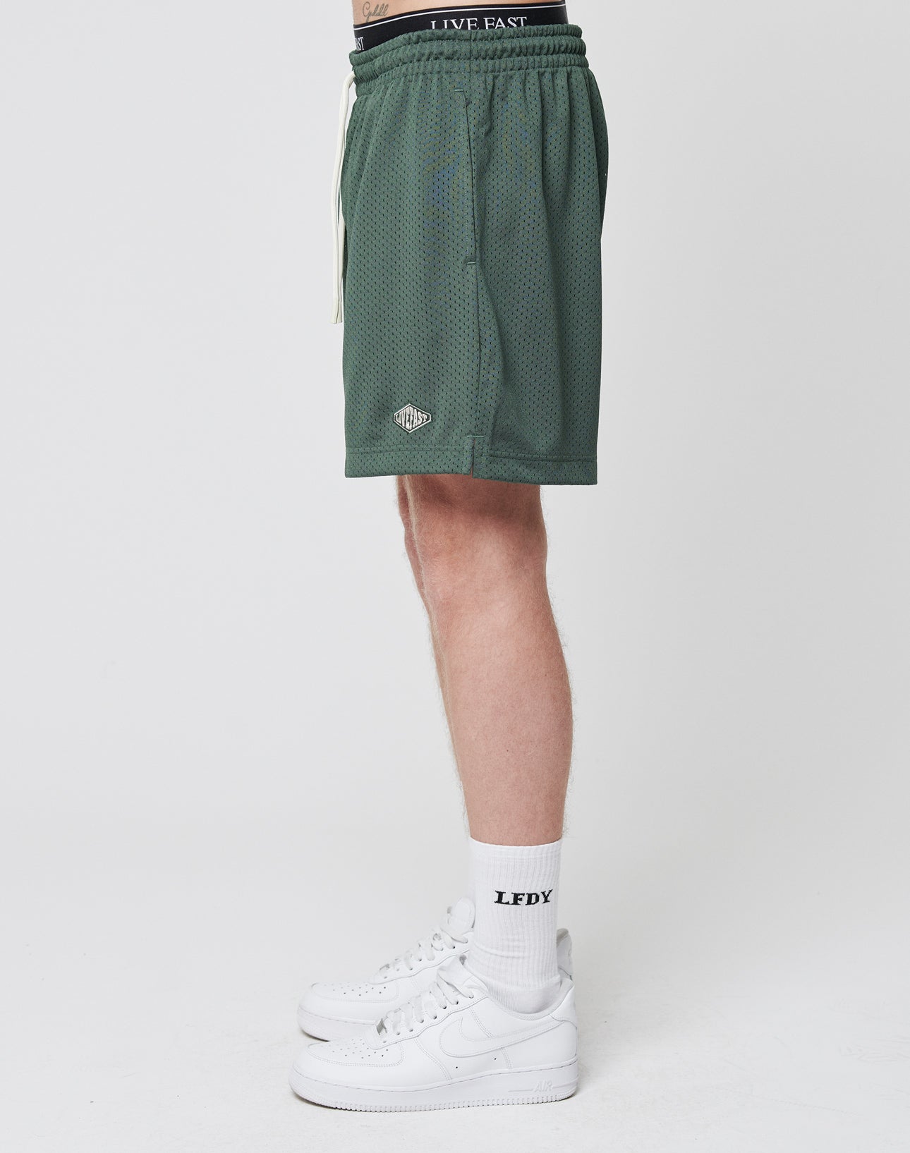 A person’s lower body is shown sideways wearing LIVE FAST DIE YOUNG Basic Mesh Shorts in green with an elastic waistband and logo patch, paired with white LFDY socks and white Nike Air Force 1 sneakers against a plain white background.