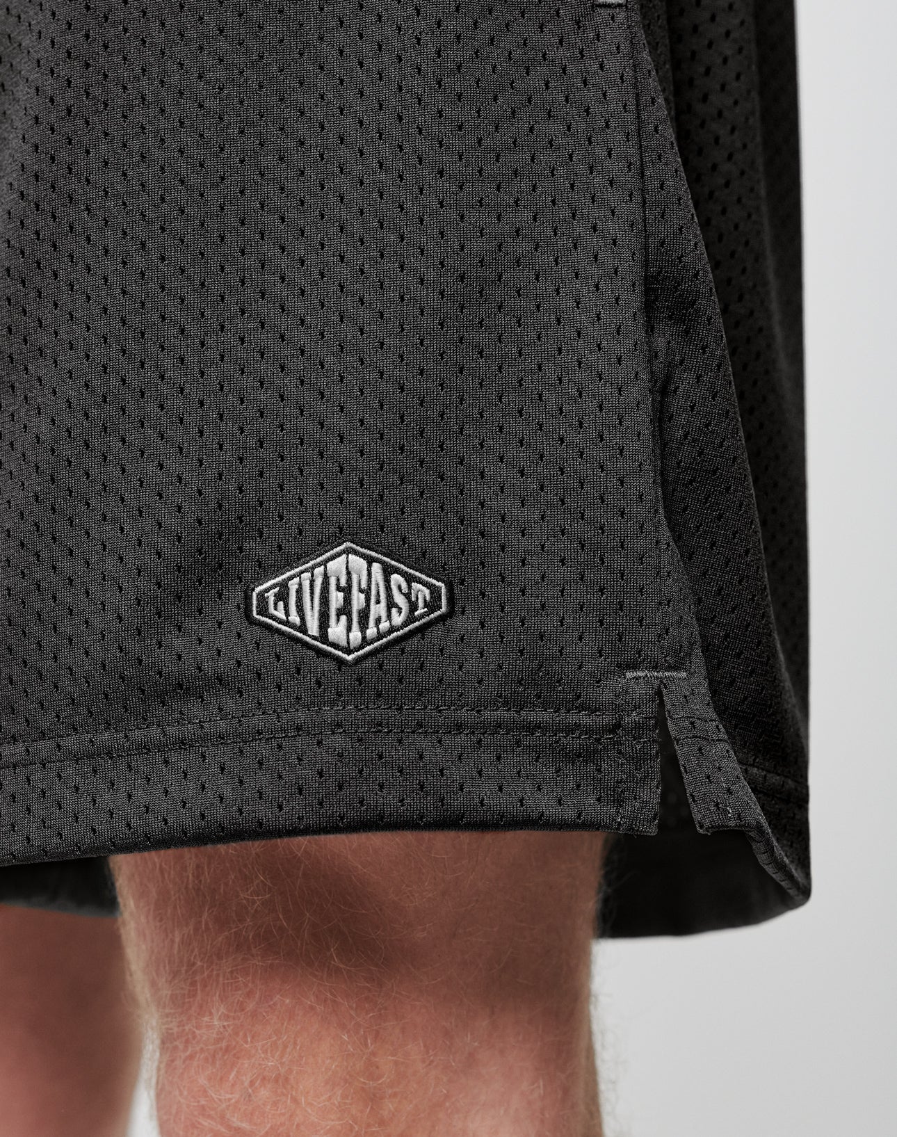 Close-up of the lower part of LIVE FAST DIE YOUNG Basic Mesh Shorts in black, featuring an elastic waistband and a white LIVE FAST logo patch on the hem, made from 100% polyester and worn by a light-skinned person.
