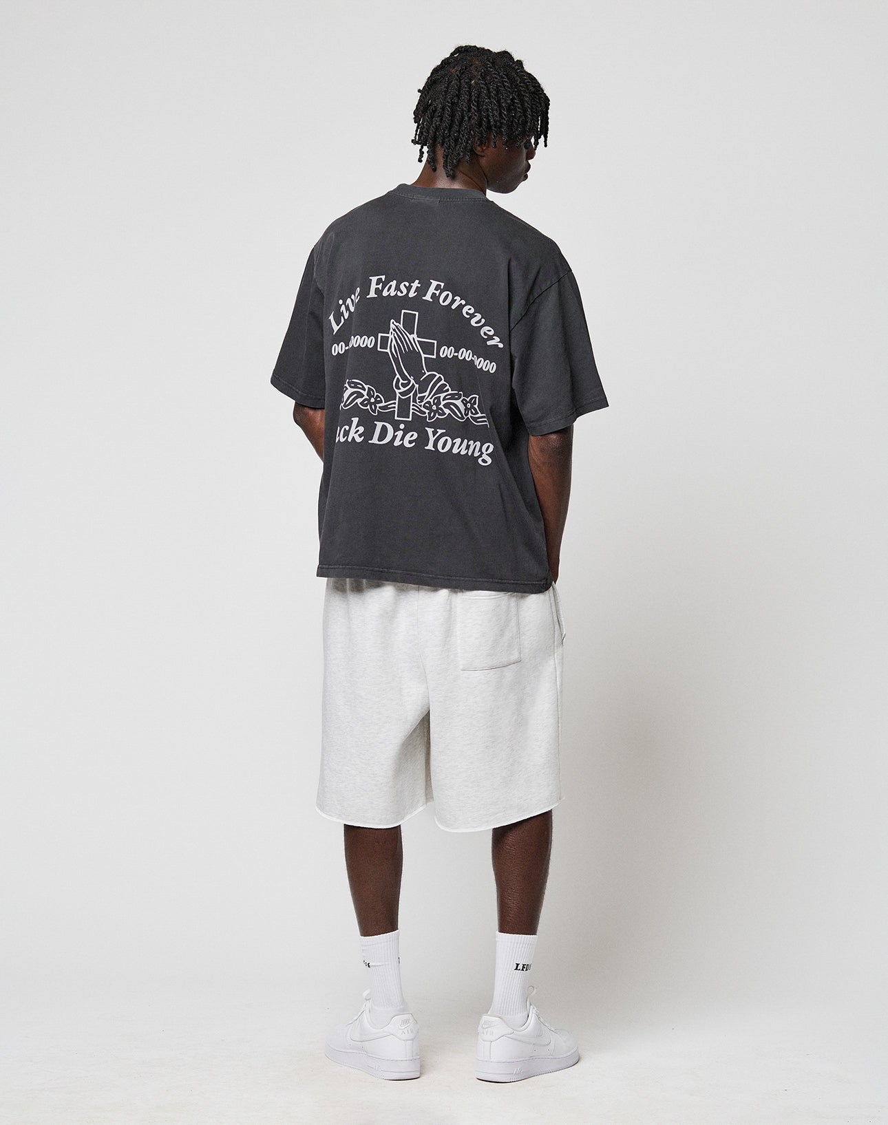 A person faces away, wearing the LIVE FAST DIE YOUNG LFDY Praying Tee with white Liv Fast Forever, Luck Die Young front and back print, light gray shorts, white socks, and white sneakers against a plain background.