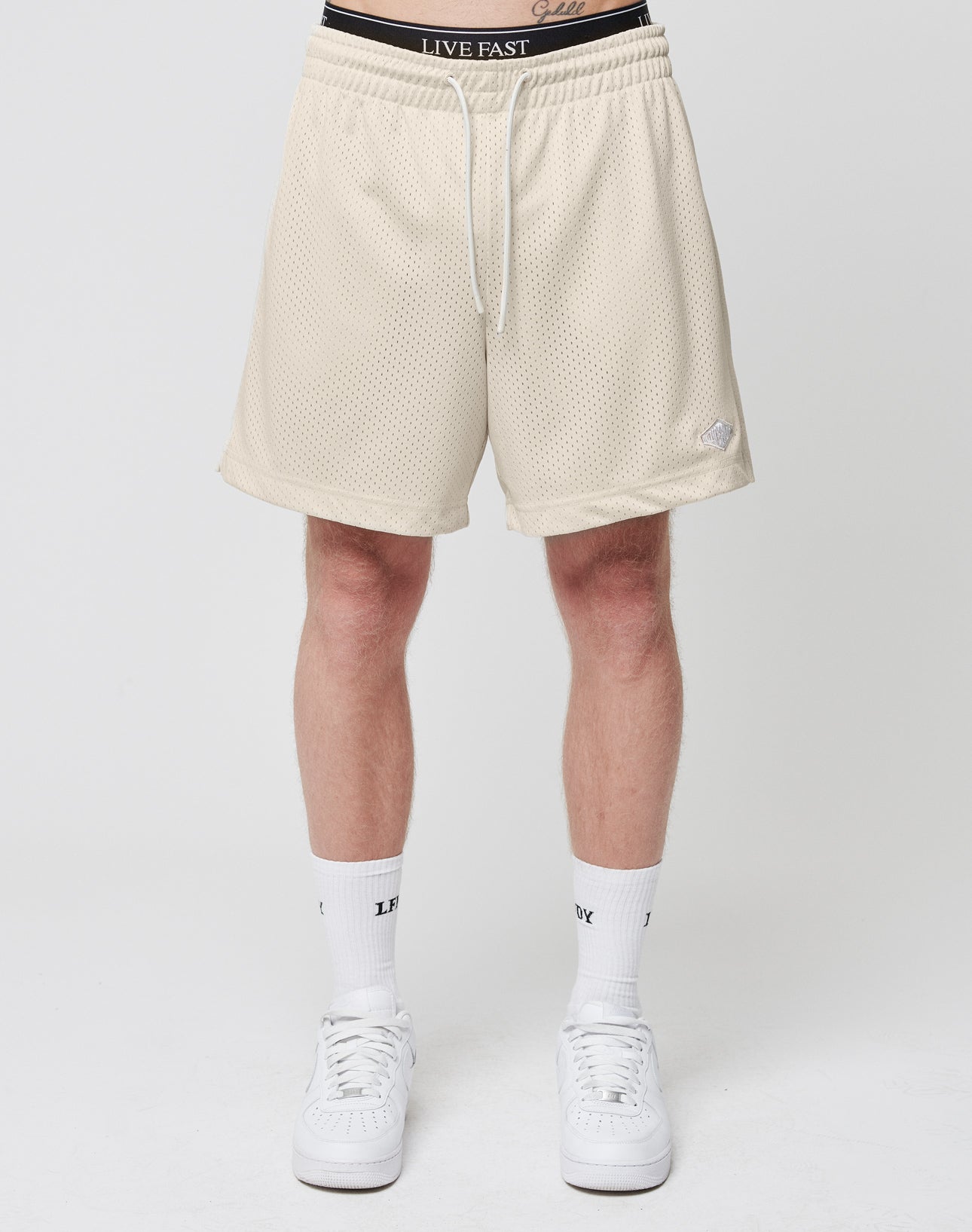 A person wears LIVE FAST DIE YOUNG Basic Mesh Shorts in light beige, white LIV-branded socks, and white sneakers, standing against a plain white background with only their lower half visible.