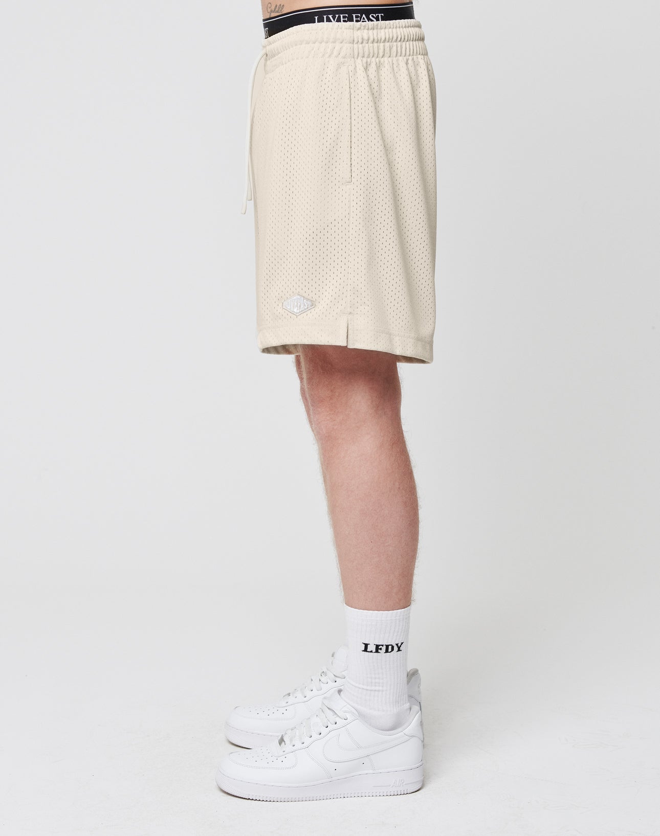 A person, shown from the waist down, models LIVE FAST DIE YOUNG Basic Mesh Shorts in beige with a logo patch, paired with white LFDY crew socks and white Nike sneakers against a plain white background.