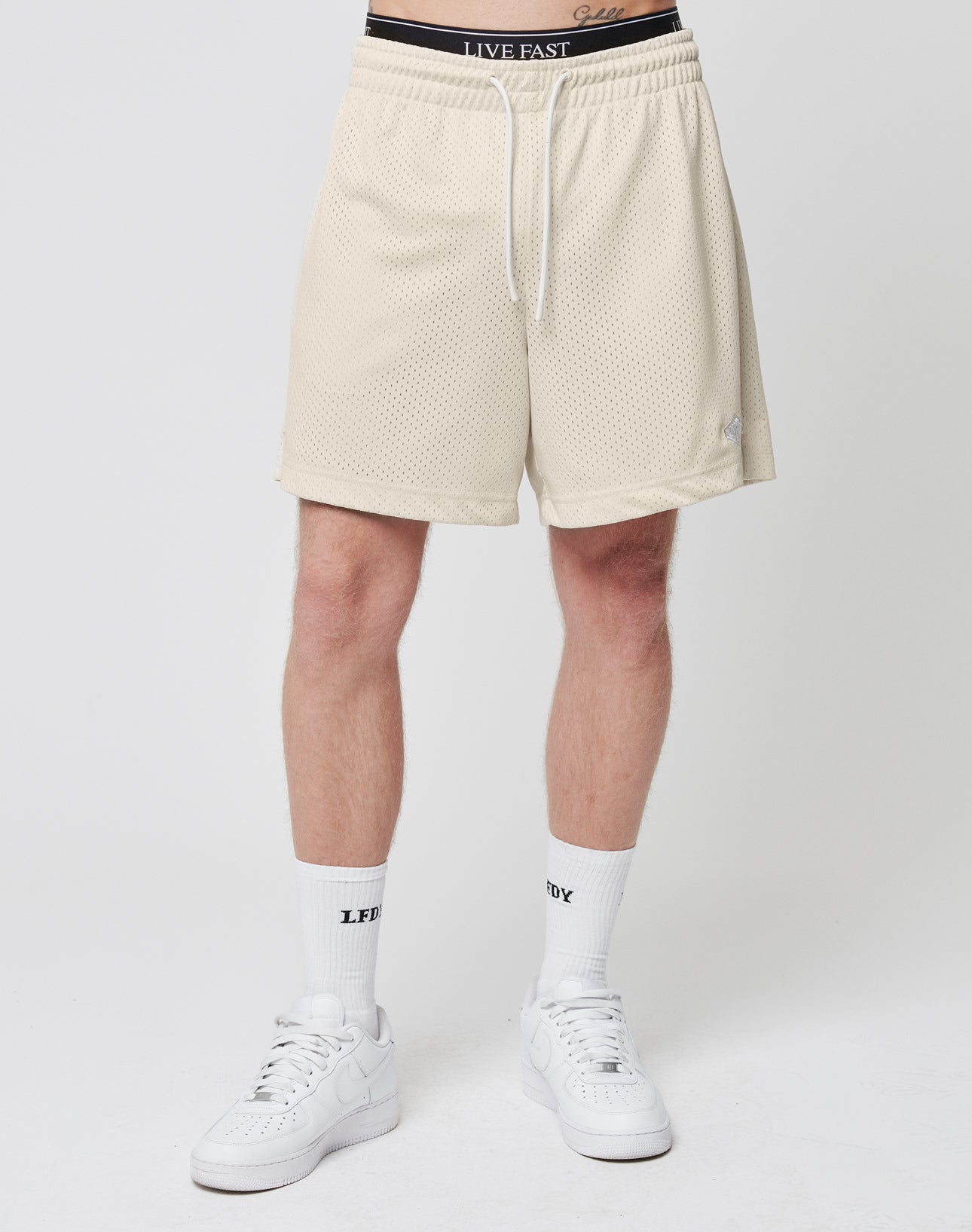 Someone wearing LIVE FAST DIE YOUNG Basic Mesh Shorts in beige, featuring a logo patch; paired with white LIVE FAST waistband underwear, white socks with LFTV text, and white sneakers, shown on a plain white background. Regular fit for comfort.