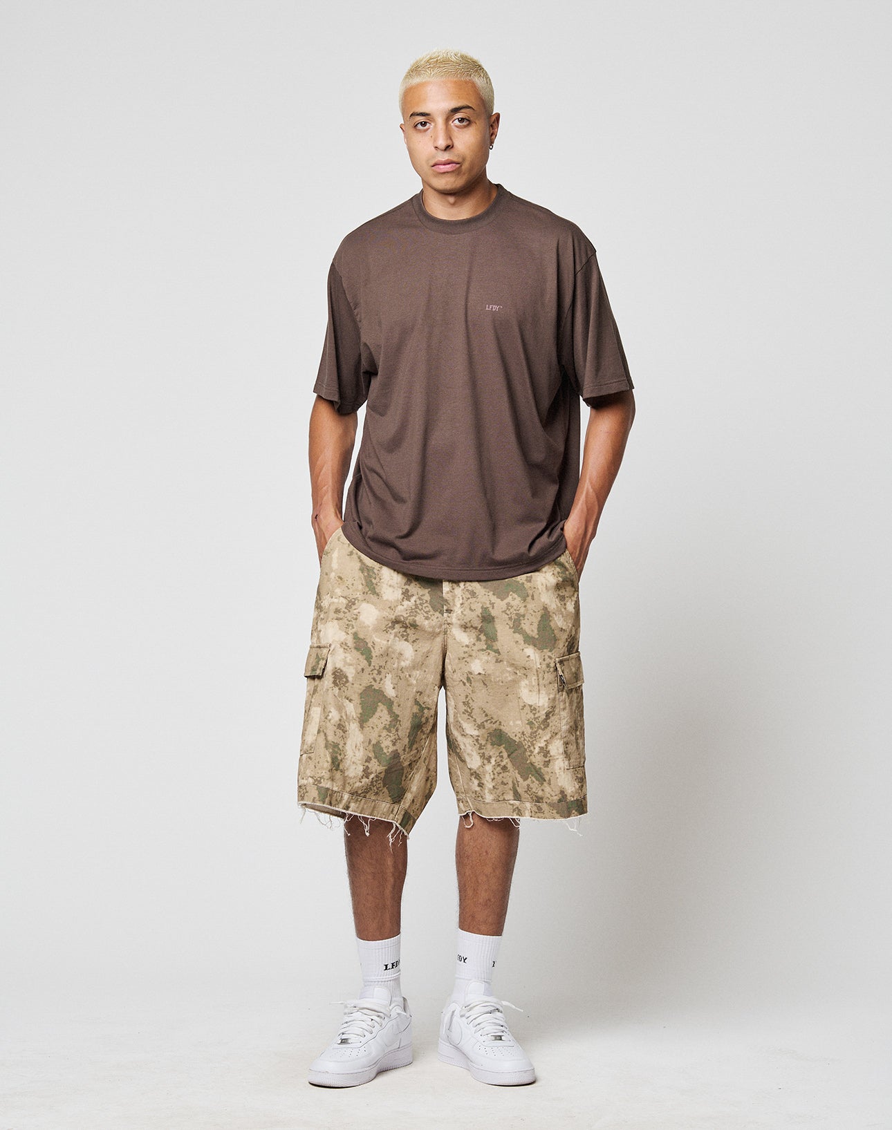 A person with short blonde hair wears a loose brown T-shirt, LIVE FAST DIE YOUNG LFDY Cargo Shorts in tan camouflage, white socks, and white sneakers, standing with hands in pockets against a plain light background.