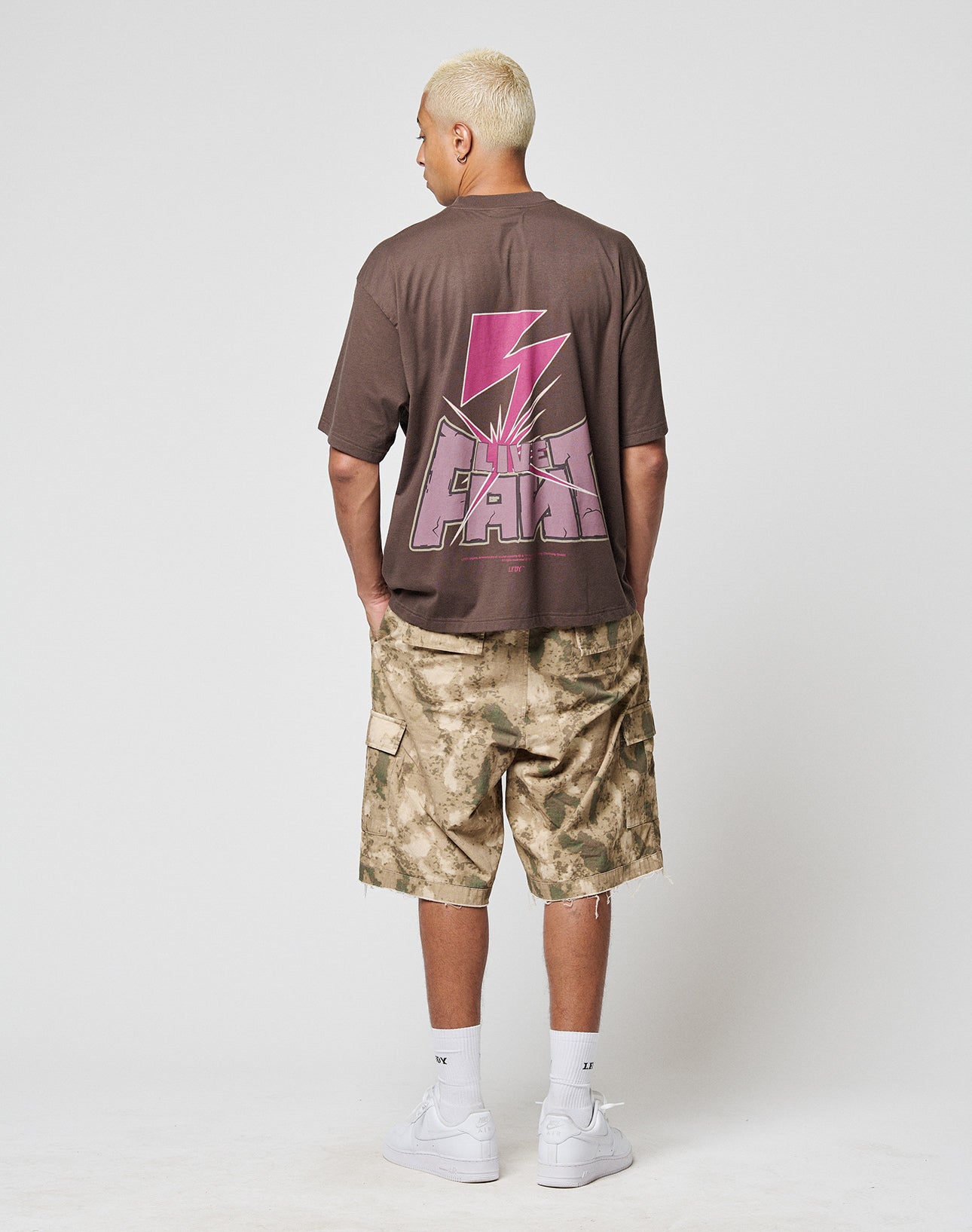A person with short blonde hair faces away, wearing the oversized LIVE FAST DIE YOUNG LFDY Flash Tee featuring a pink lightning bolt and LIFE FAST back print, camouflage cargo shorts, white socks, and white sneakers.