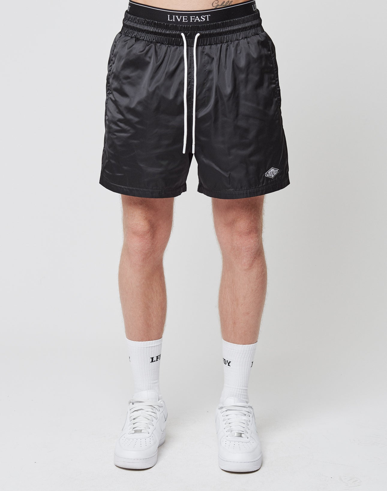 A person in LIVE FAST DIE YOUNG Basic Satin Shorts—shiny black, with an elastic waistband and logo patch—pairs them with white socks and sneakers, posing against a plain white background.