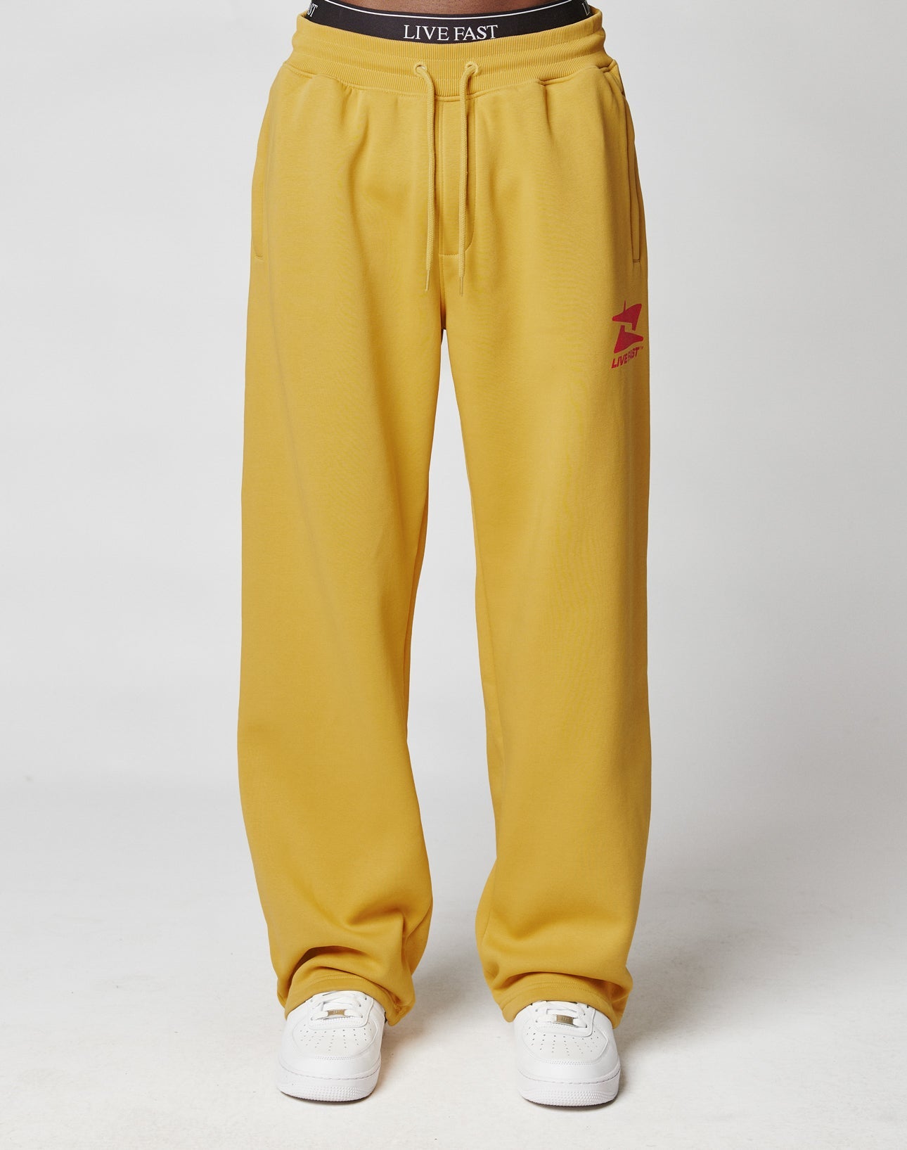 A person wears the LIVE FAST DIE YOUNG Live Fast Sweatsuit Jogger in mustard yellow with an oversize fit, elastic waistband, red logo on the left thigh, white sneakers, and a black LIVE FAST waistband against a plain white background.