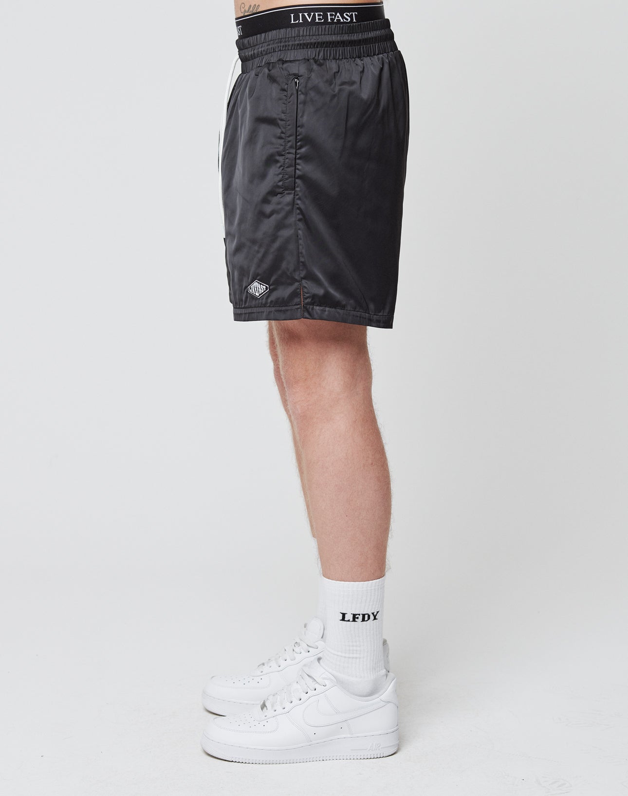 A person stands sideways wearing LIVE FAST DIE YOUNG Basic Satin Shorts featuring a logo patch and “LIVE FAST” on the elastic waistband, paired with white “LFDY” socks and white sneakers, against a plain white background.