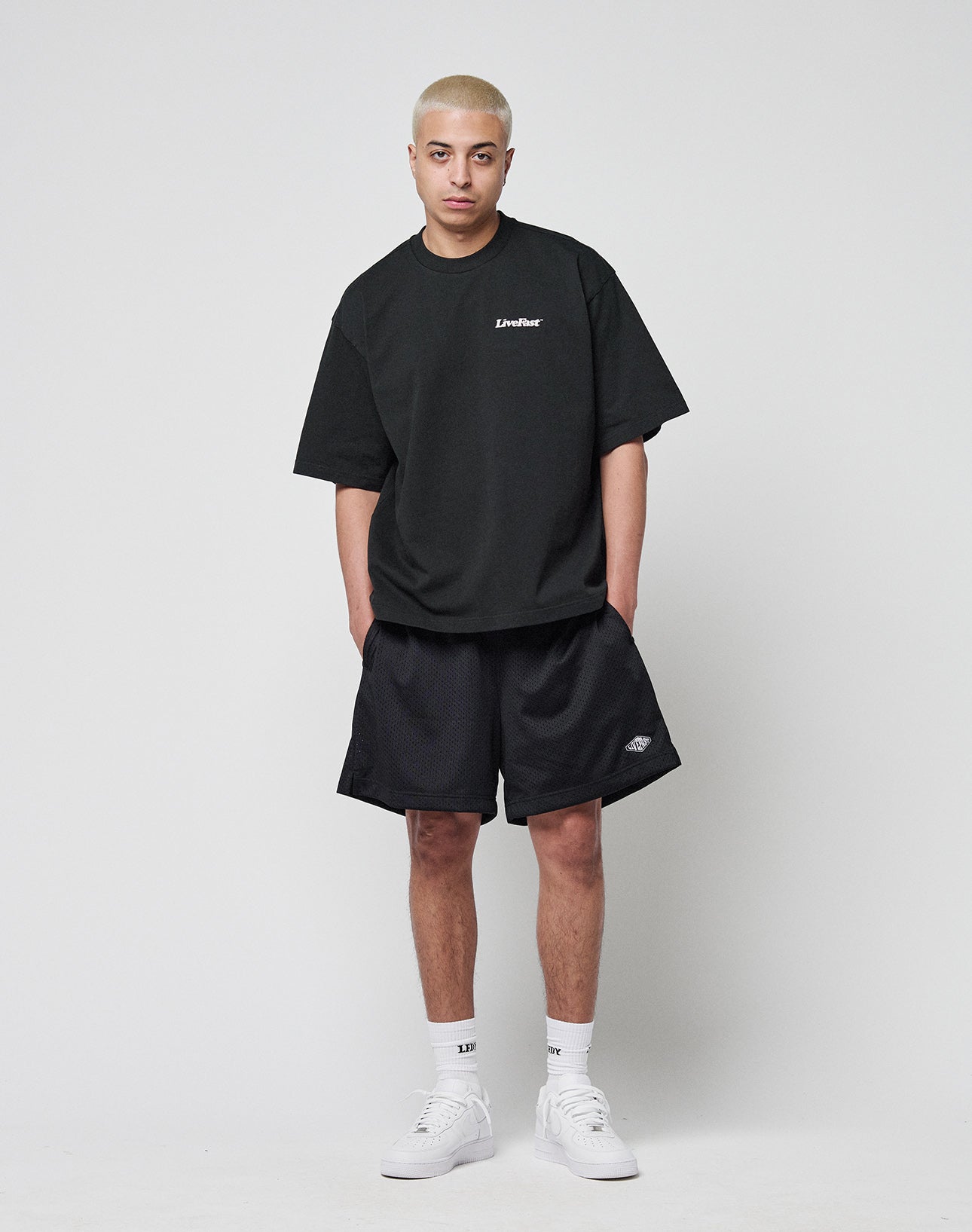 A person with short blond hair wears a LIVE FAST DIE YOUNG Basic 280 Tee in black with front and back print, black shorts, white socks, and white sneakers, standing against a plain light gray background facing the camera.