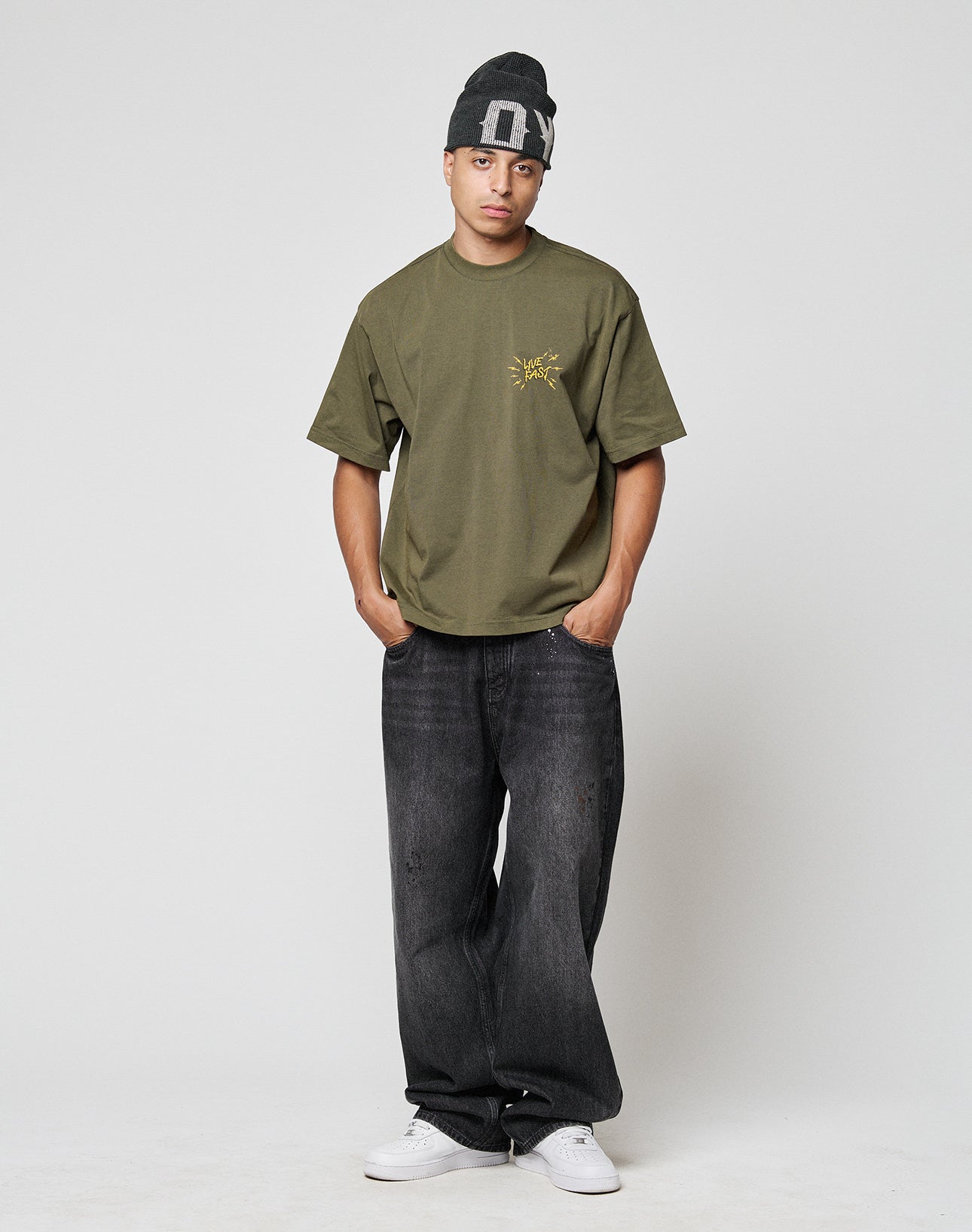 A person stands against a plain background, wearing the LIVE FAST DIE YOUNG LFDY Against us Tee in olive green with a small yellow graphic, paired with a dark gray beanie, loose black jeans, and white sneakers.