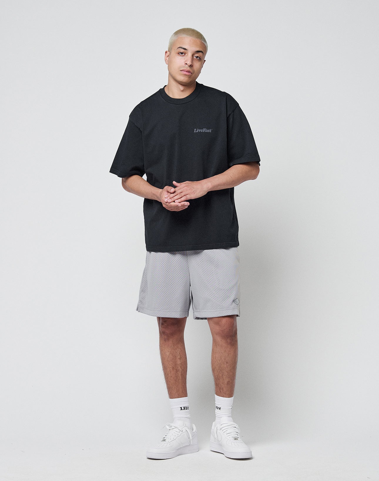 A person with short blond hair wears the LIVE FAST DIE YOUNG Basic 280 Tee in black, light gray shorts, white socks, and white sneakers, standing against a plain white background with hands clasped.
