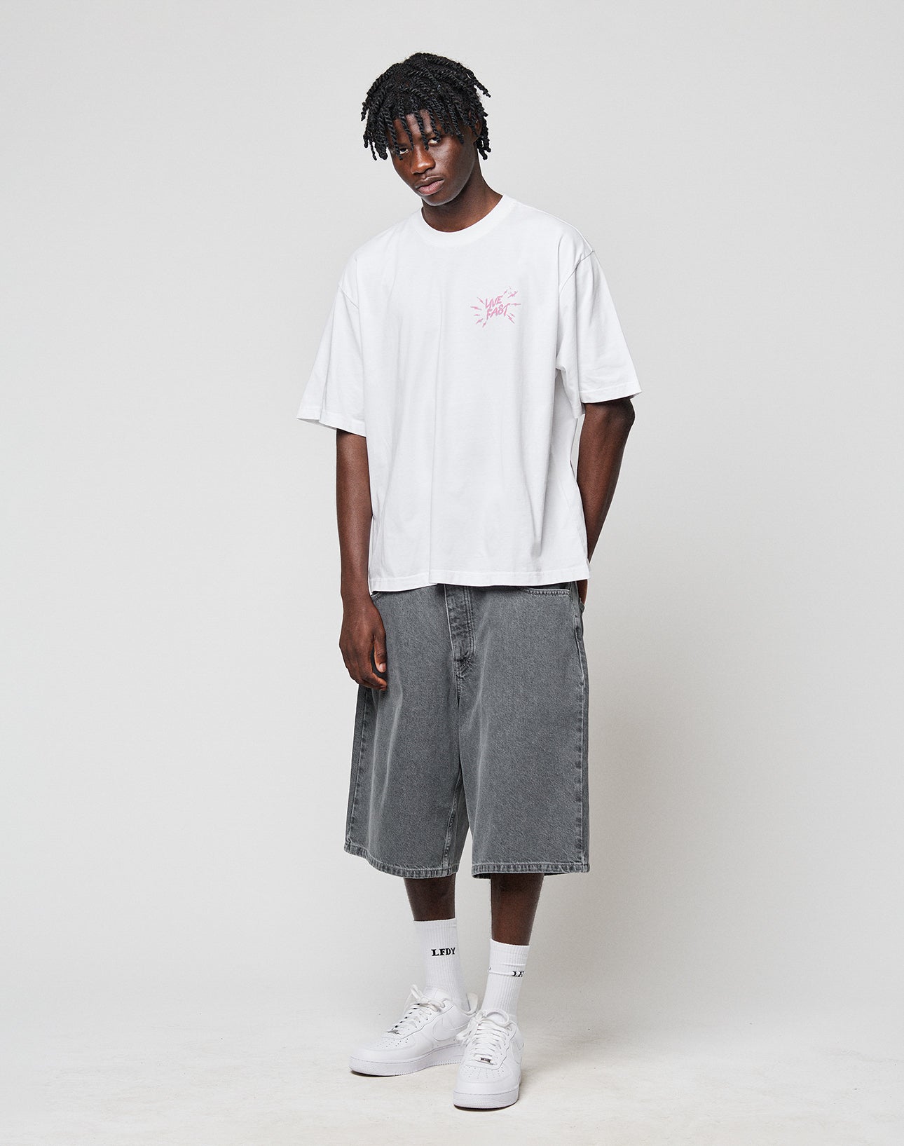 A man stands against a plain background wearing the LFDY Against us Tee by LIVE FAST DIE YOUNG—an oversize 100% cotton white T-shirt with a pink frontprint—paired with gray denim shorts, white socks, and sneakers. He looks relaxed, hands at his sides.