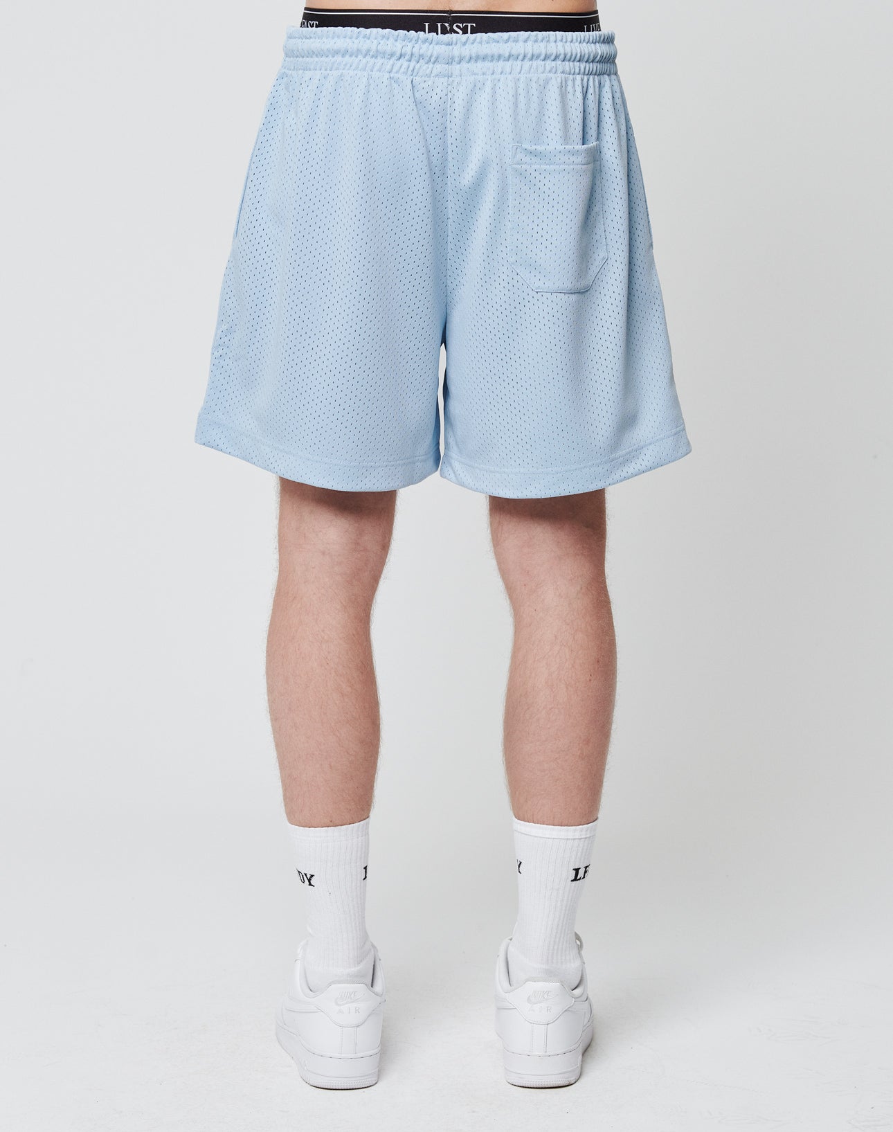A person stands facing away, wearing LIVE FAST DIE YOUNG Basic Mesh Shorts in light blue. These regular fit, 100% polyester shorts have an elastic waistband and a back pocket. Only the lower body is visible against a plain white background.
