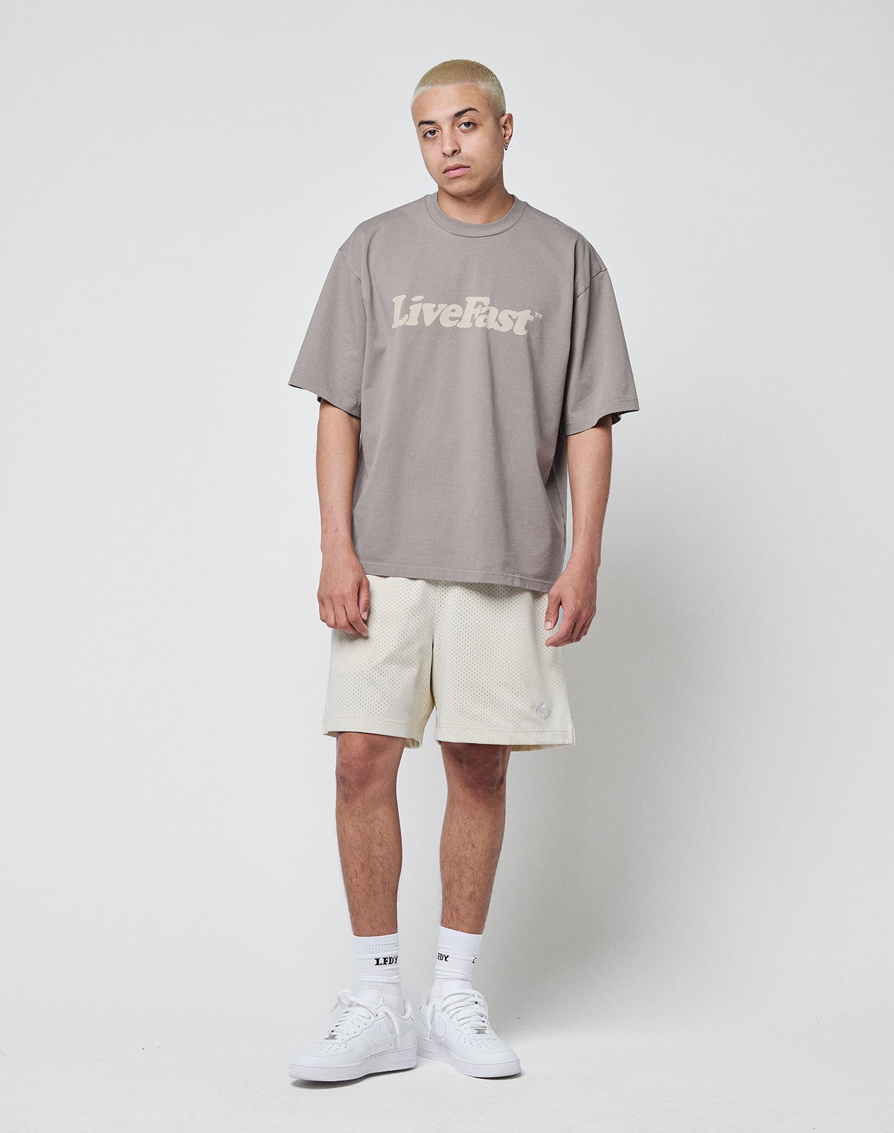 A person with short bleached hair stands against a plain white background, wearing a loose taupe LIVE FAST DIE YOUNG Basic Tee in Oversize Fit, cream mesh shorts, white socks, and white sneakers.