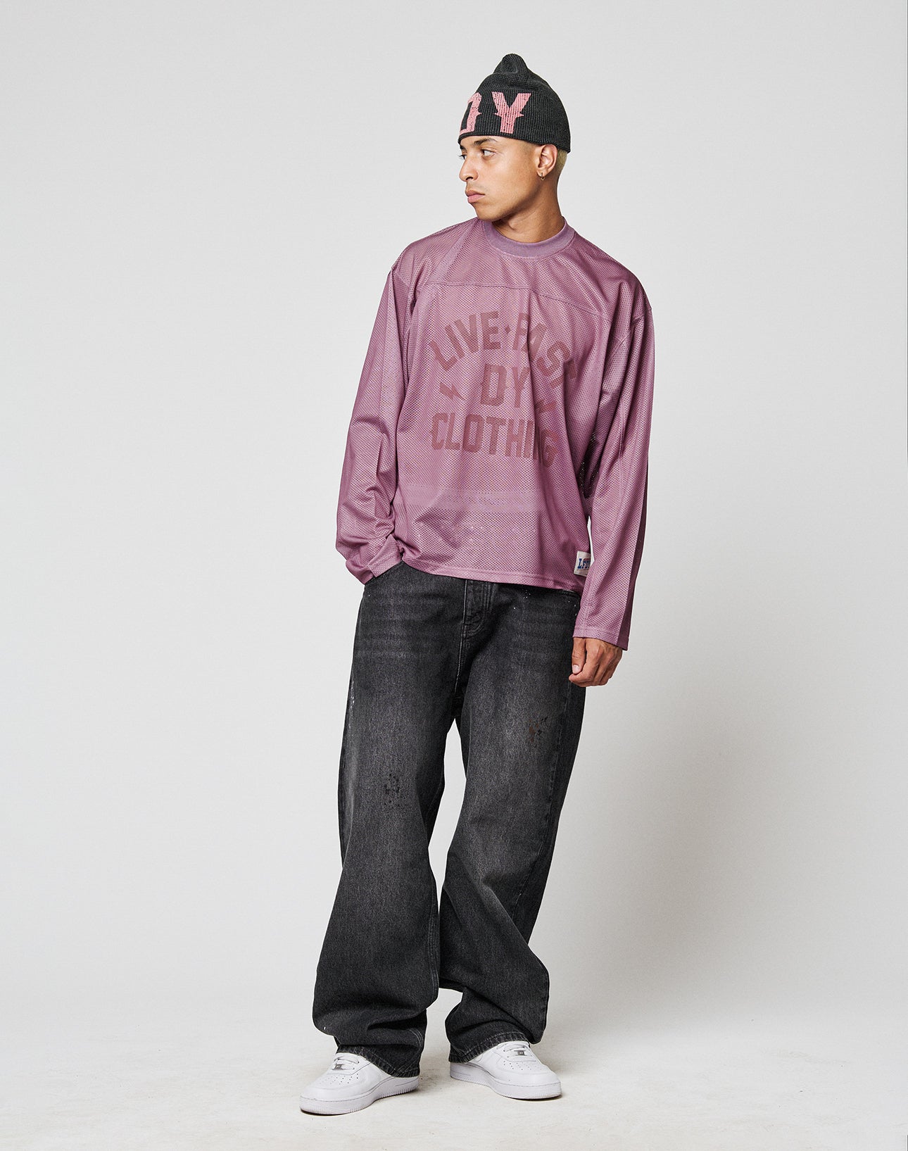 A man stands against a plain light gray background wearing a black beanie, baggy dark jeans, white sneakers, and an oversize mauve LIVE FAST DIE YOUNG LFDY Flash Jersey.