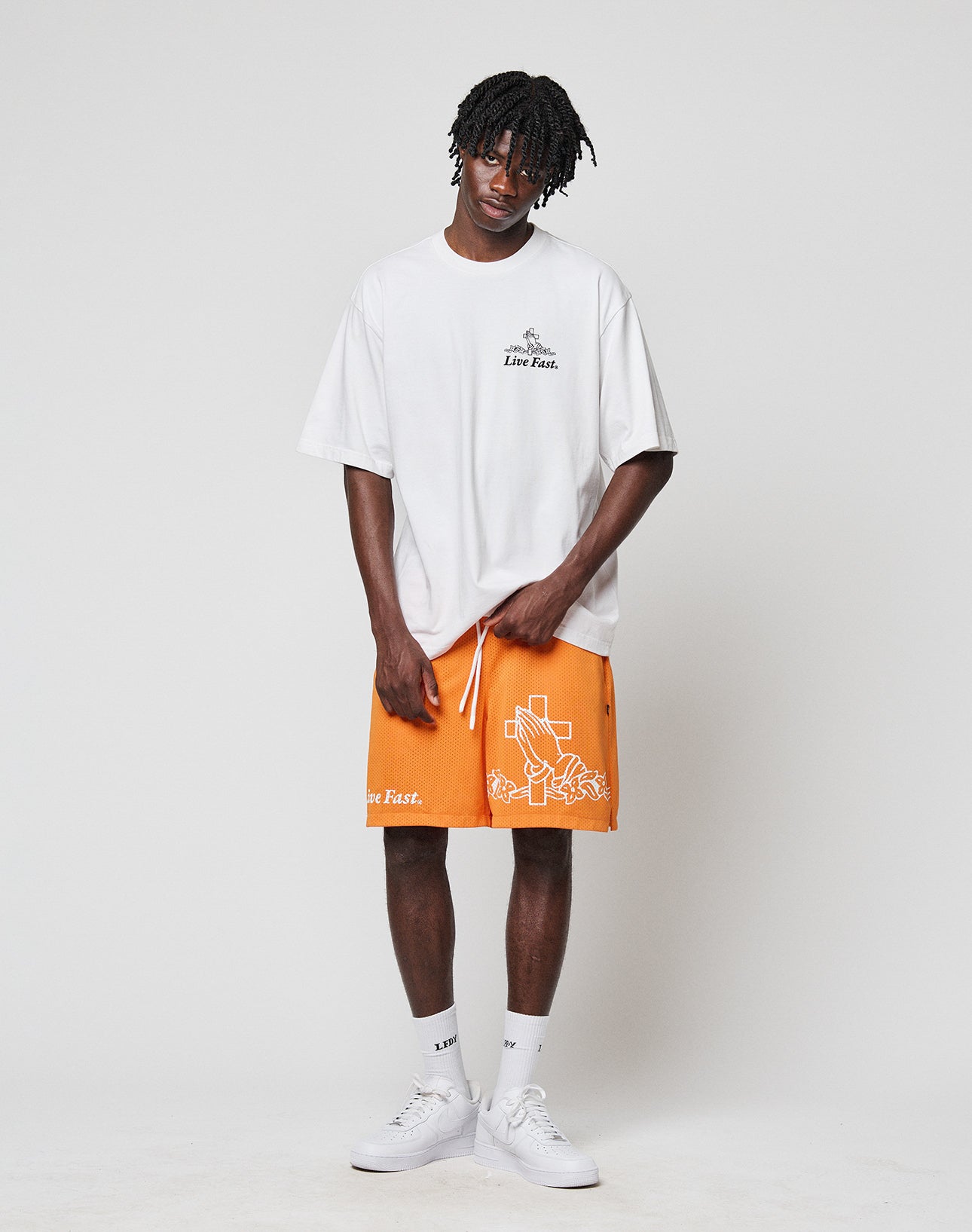 A person with short, styled hair stands in a relaxed pose against a white background, wearing the LIVE FAST DIE YOUNG LFDY Praying Tee, orange shorts with white graphics, white socks, and sneakers.