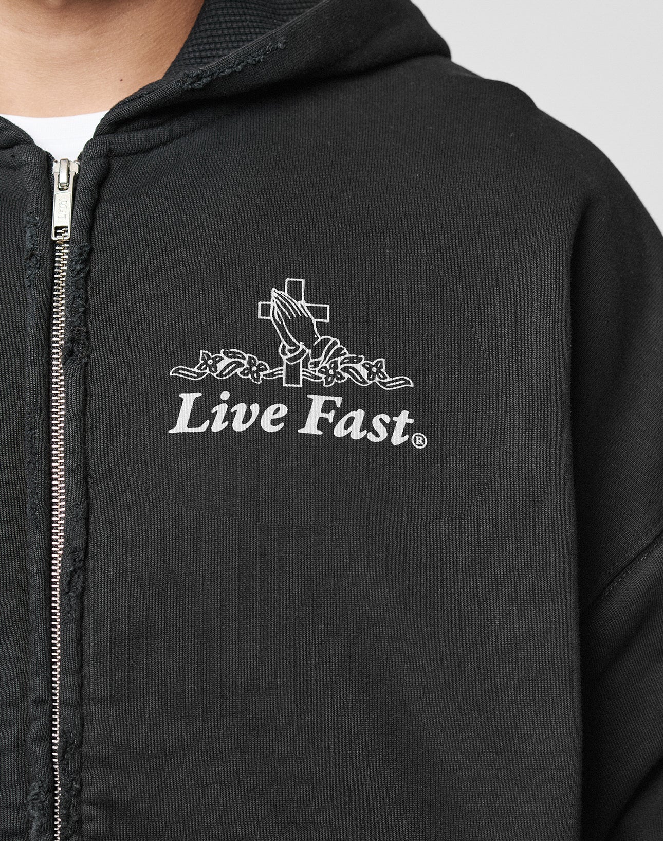 A person wears the LFDY Praying Hooded Zip by LIVE FAST DIE YOUNG, an oversized black zip-up hoodie with a white graphic of praying hands, a cross, and Live Fast on the chest. Features subtle distress-effect details for an edgy look.