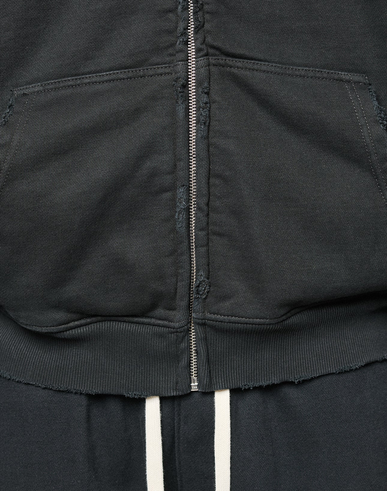 Close-up of the LFDY Praying Hooded Zip by LIVE FAST DIE YOUNG, a black oversized hoodie with a metal zipper, front pouch pocket, frayed hem Distress-Effekt, paired with dark pants featuring two vertical white stripes.