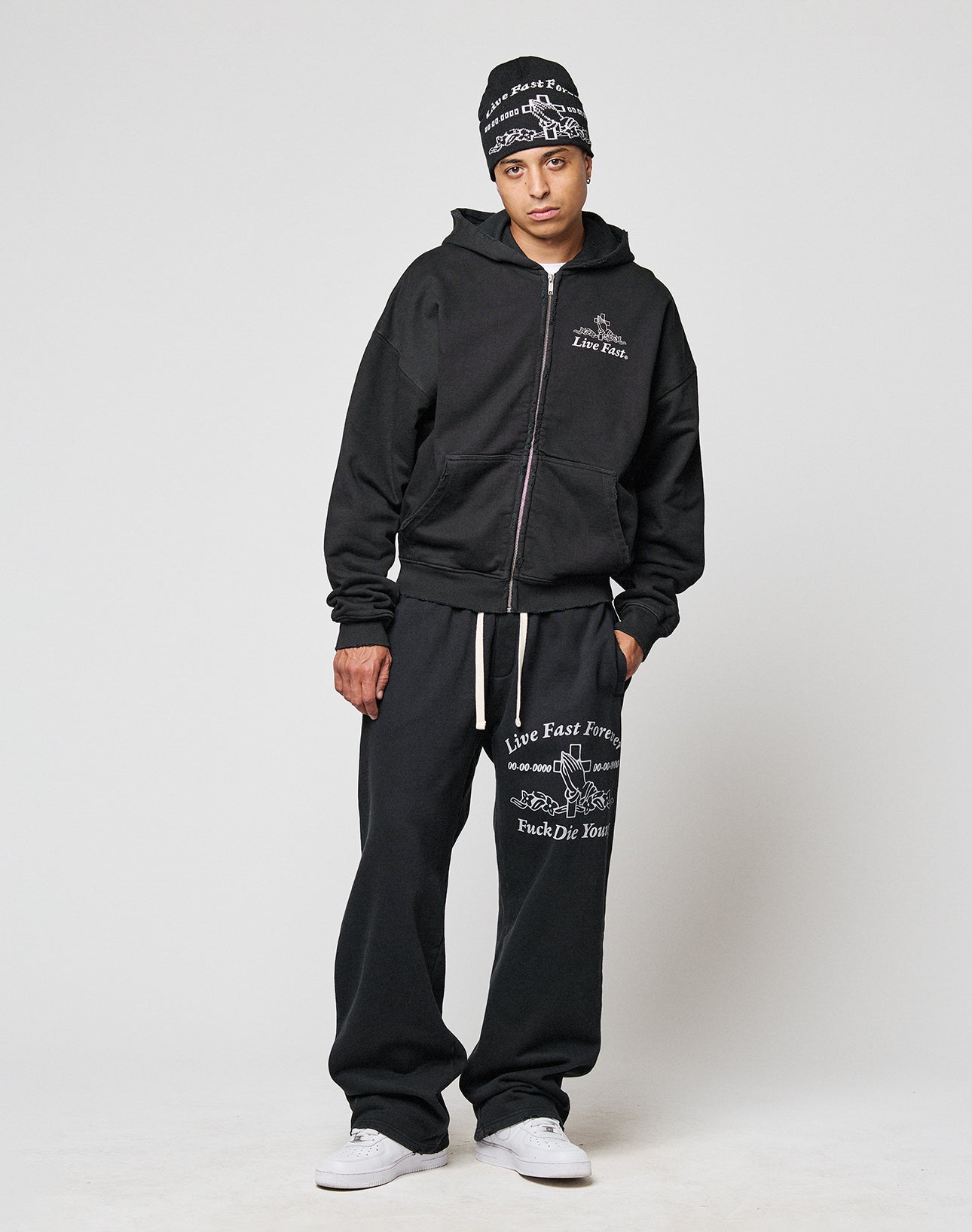 A person stands against a plain background wearing an oversize black beanie, black hoodie, and LIVE FAST DIE YOUNG LFDY Praying Jogger sweatpants with white graphics, text, a comfy Elastikbund, one hand in their pocket, and white sneakers.