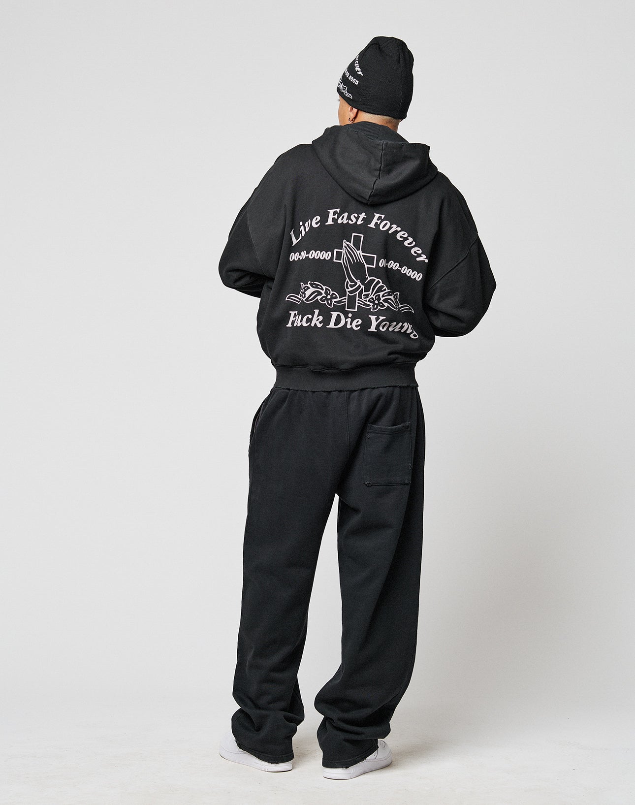 Facing away, a person wears the LFDY Praying Hooded Zip by LIVE FAST DIE YOUNG with bold graphics, sweatpants, beanie, and white sneakers. The hoodie’s distressed effect adds extra edge.
