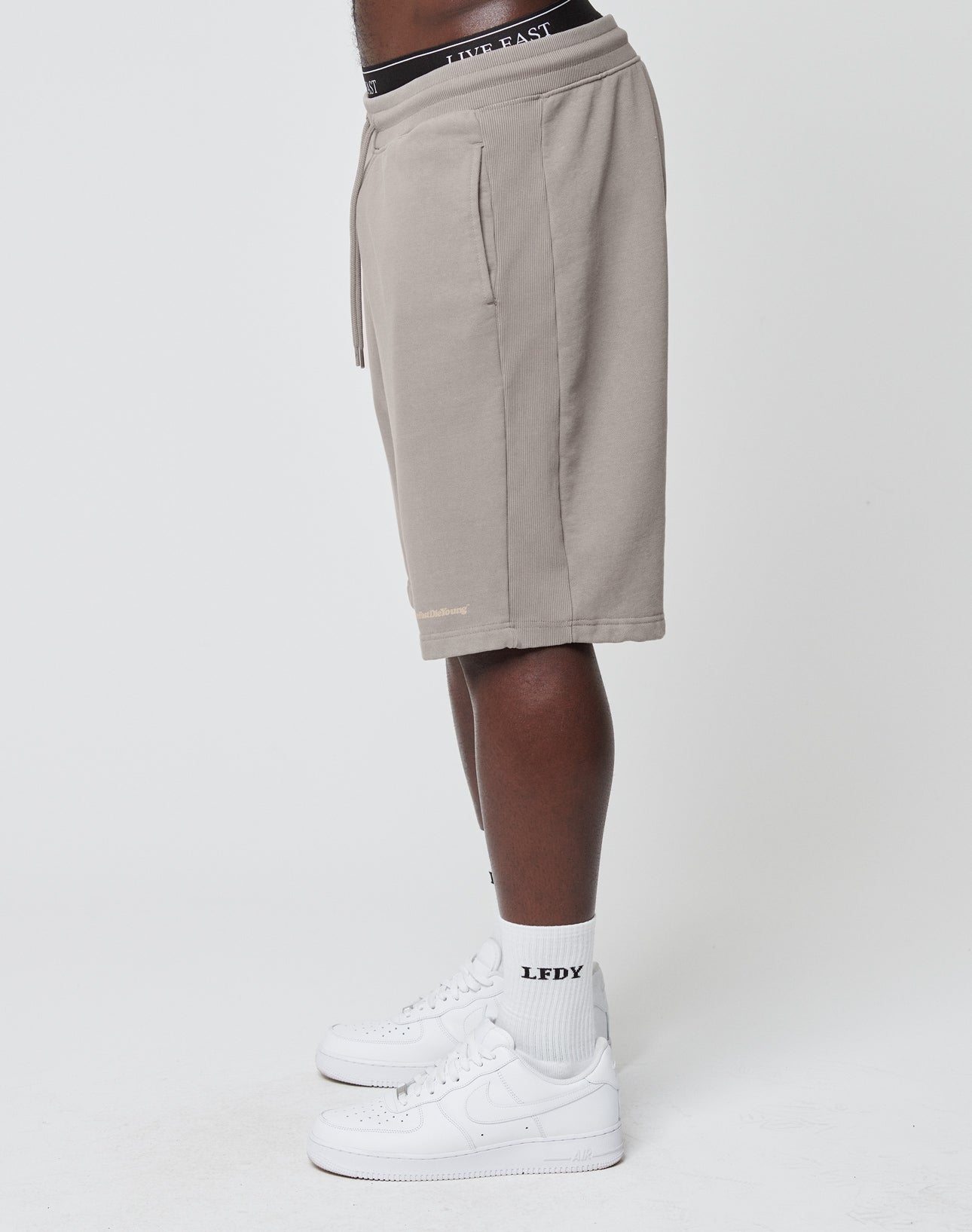 A person stands sideways in taupe Basic Heavy Shorts by LIVE FAST DIE YOUNG with a Regular Fit, black waistband, paired with white LFDY socks and white Nike sneakers against a plain white background.
