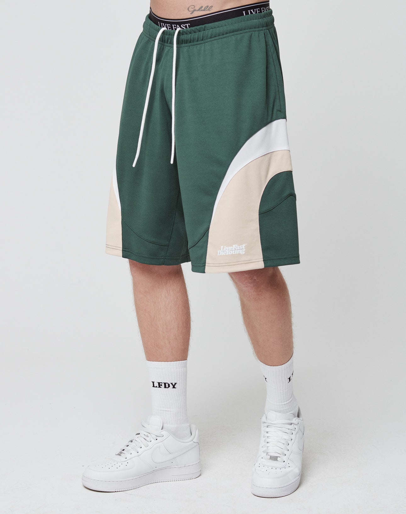 A person in green, white, and beige LIVE FAST DIE YOUNG Basic Trackshorts with a logo-print waistband, white LFDY socks, and white sneakers stands against a plain light background.