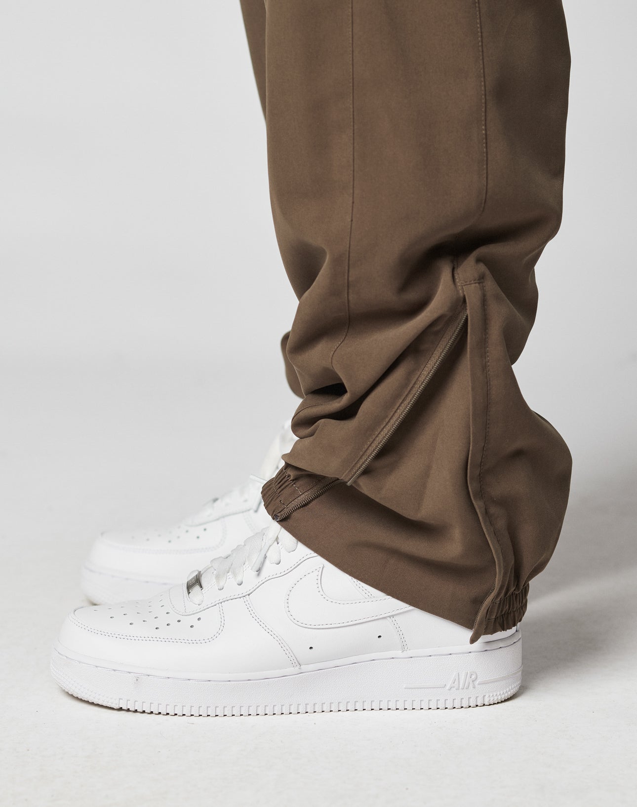 A close-up side view of a person wearing LIVE FAST DIE YOUNG LFDY Trackpants with zippered hems and elastic waistband, styled with white Nike Air Force 1s against a plain background.
