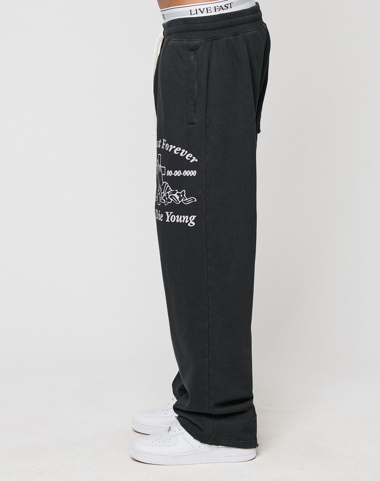 A person stands in profile against a white background wearing the LFDY Praying Jogger by LIVE FAST DIE YOUNG, featuring black sweatpants with white graphics, LIVE FAST elastic waistband, oversize fit, and white sneakers.