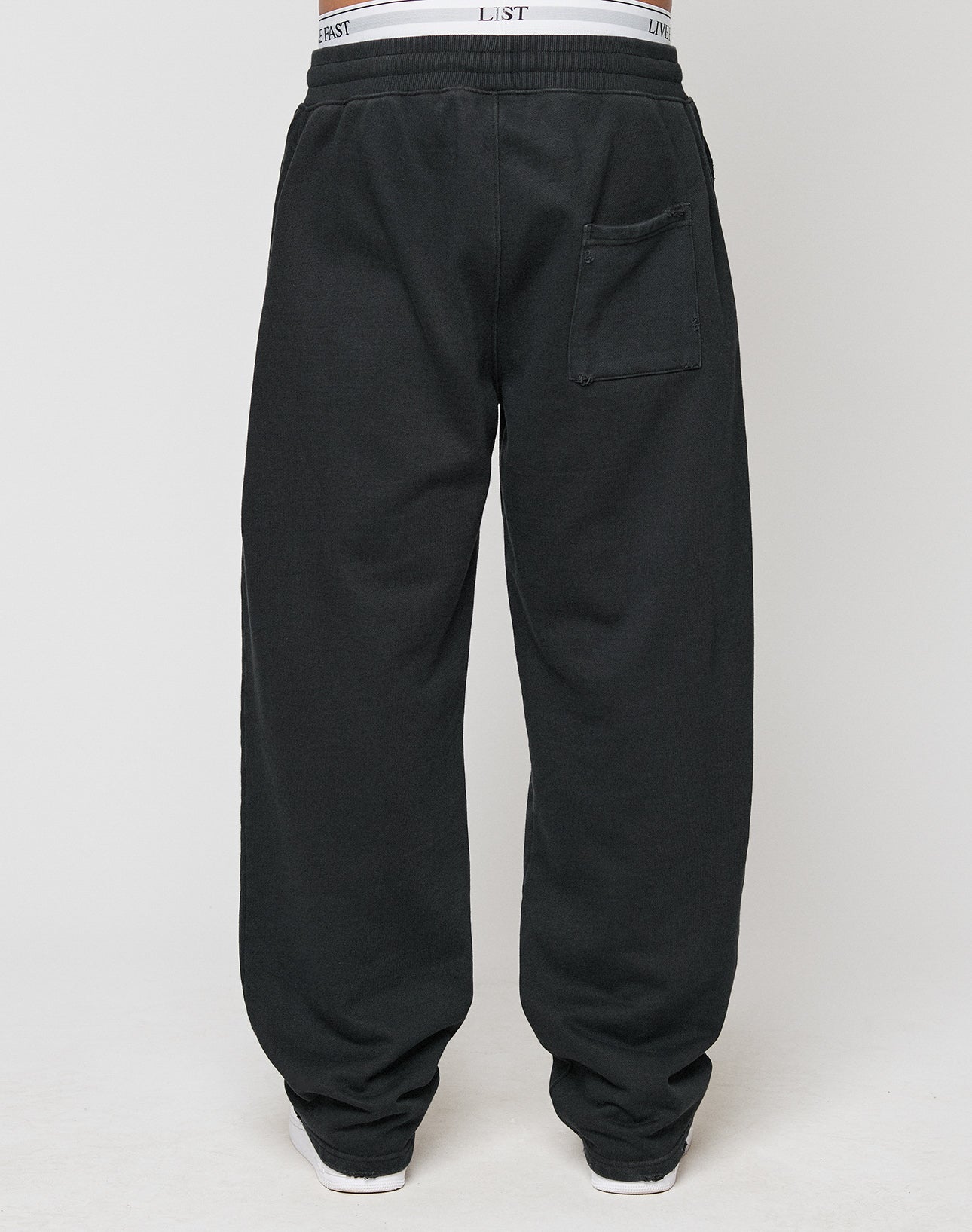 A person is seen from the back wearing LIVE FAST DIE YOUNG’s LFDY Praying Jogger—loose-fitting black sweatpants with an oversize fit, a visible back pocket, and white underwear waistband peeking over the elastic waist.