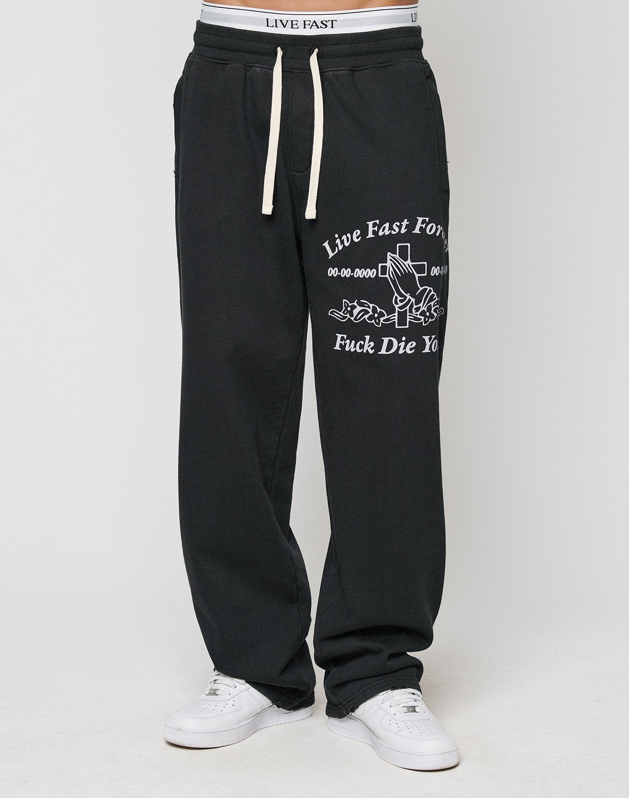 A person wears the LFDY Praying Jogger by LIVE FAST DIE YOUNG—black sweatpants with an elastic waistband reading LIVE FAST, bold white graphics, Live Fast F*** Die Yo text, paired with white sneakers and an oversized fit.