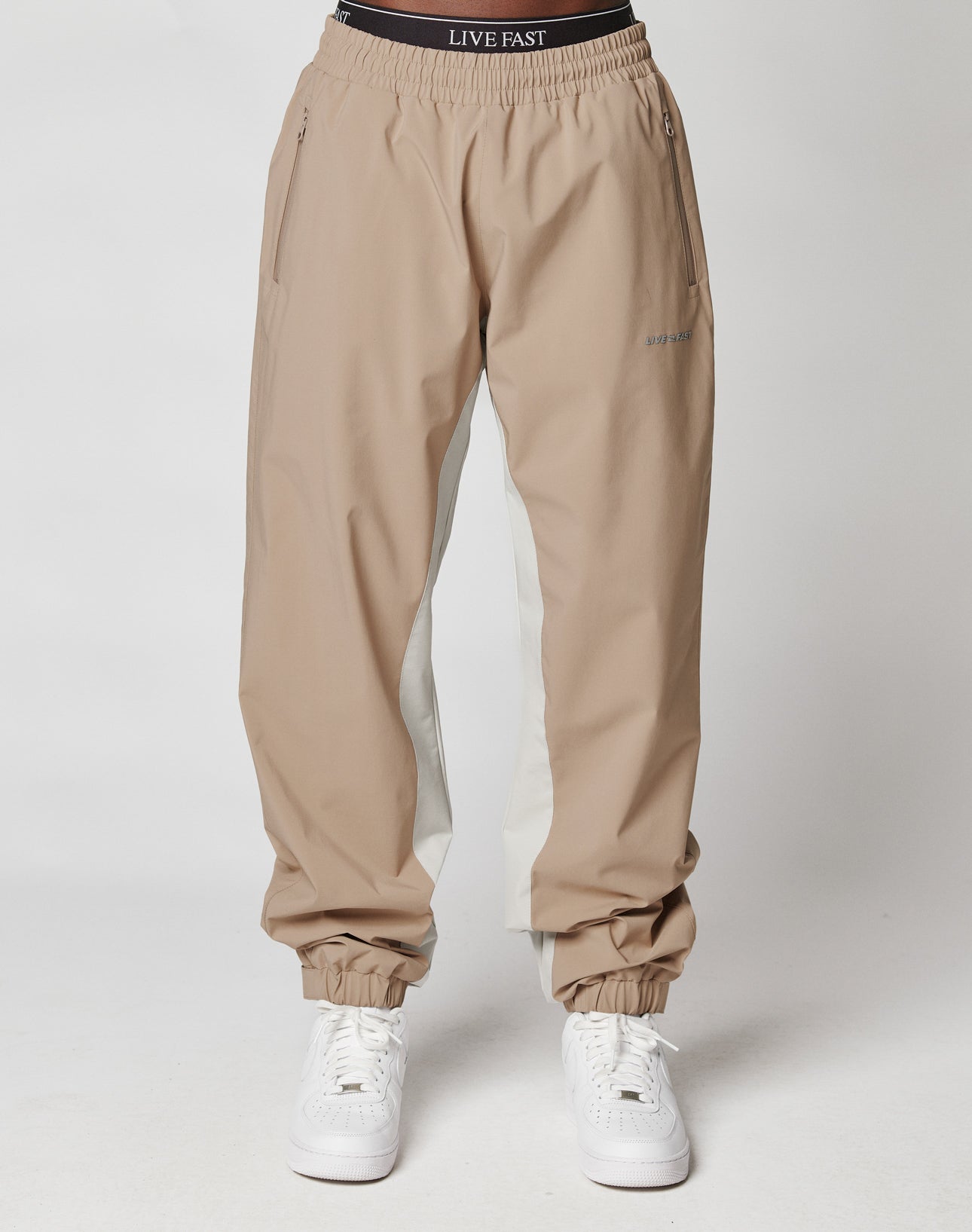 A person wears LIVE FAST DIE YOUNG Basic Shell Pants in tan and beige with an elastic drawstring waistband and cuffs, paired with white sneakers. The black waistband reads “LIVE FAST” in white, set against a plain white background.
