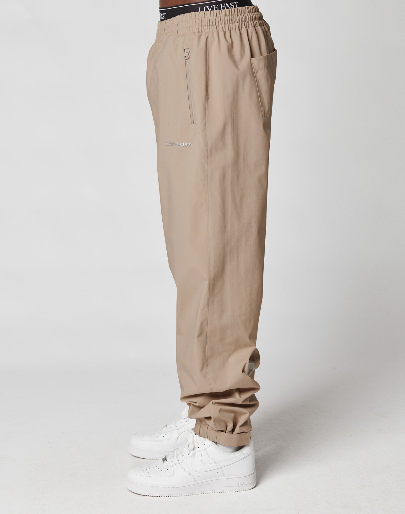 A person wearing LIVE FAST DIE YOUNG Basic Shell Pants with an elastic waistband and zippered pockets, paired with white Nike sneakers, stands in profile against a plain white background.