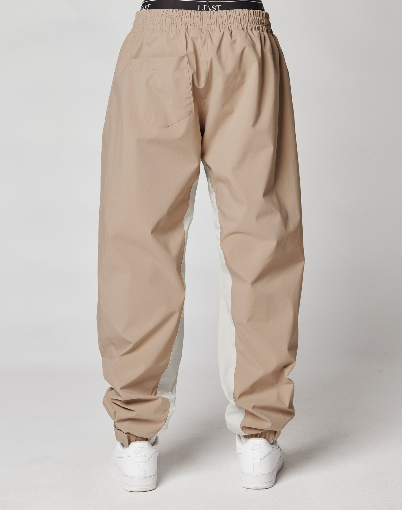 A person wearing LIVE FAST DIE YOUNG Basic Shell Pants in loose-fitting, two-tone beige and white with elastic waistband, drawcord, and cuffs, paired with white sneakers, shown from the back against a plain background.
