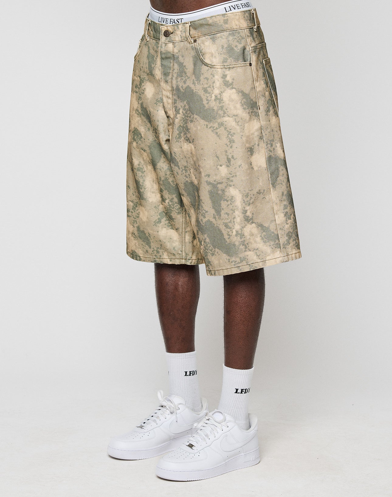 A person in LIVE FAST DIE YOUNG Canvas Shorts with a baggy fit and straight leg, white socks with LEDF text, and white Nike Air Force 1s stands against a plain white background.
