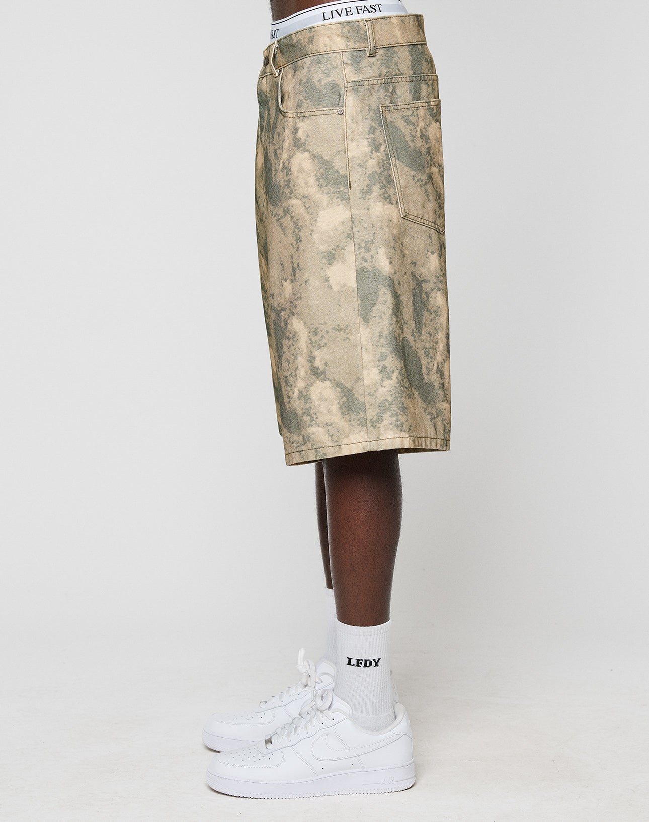 A person stands sideways in beige camouflage Canvas Shorts by LIVE FAST DIE YOUNG, paired with white LFDY waistband boxers, white LFDY socks, and white sneakers against a plain light background.