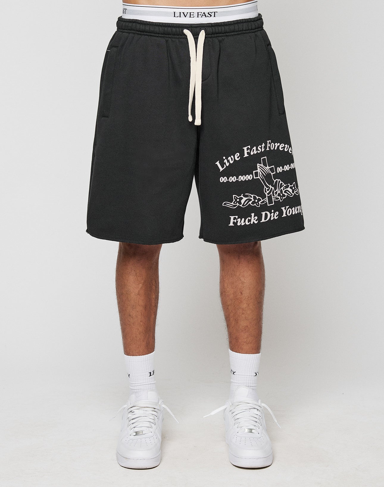 A person wears LIVE FAST DIE YOUNG LFDY Praying Shorts (regular fit, black, white Live Fast Forever and Fuck Die Young text, side pockets), white sneakers, and a visible white LIVE FAST waistband. Photo is waist-down against a plain background.