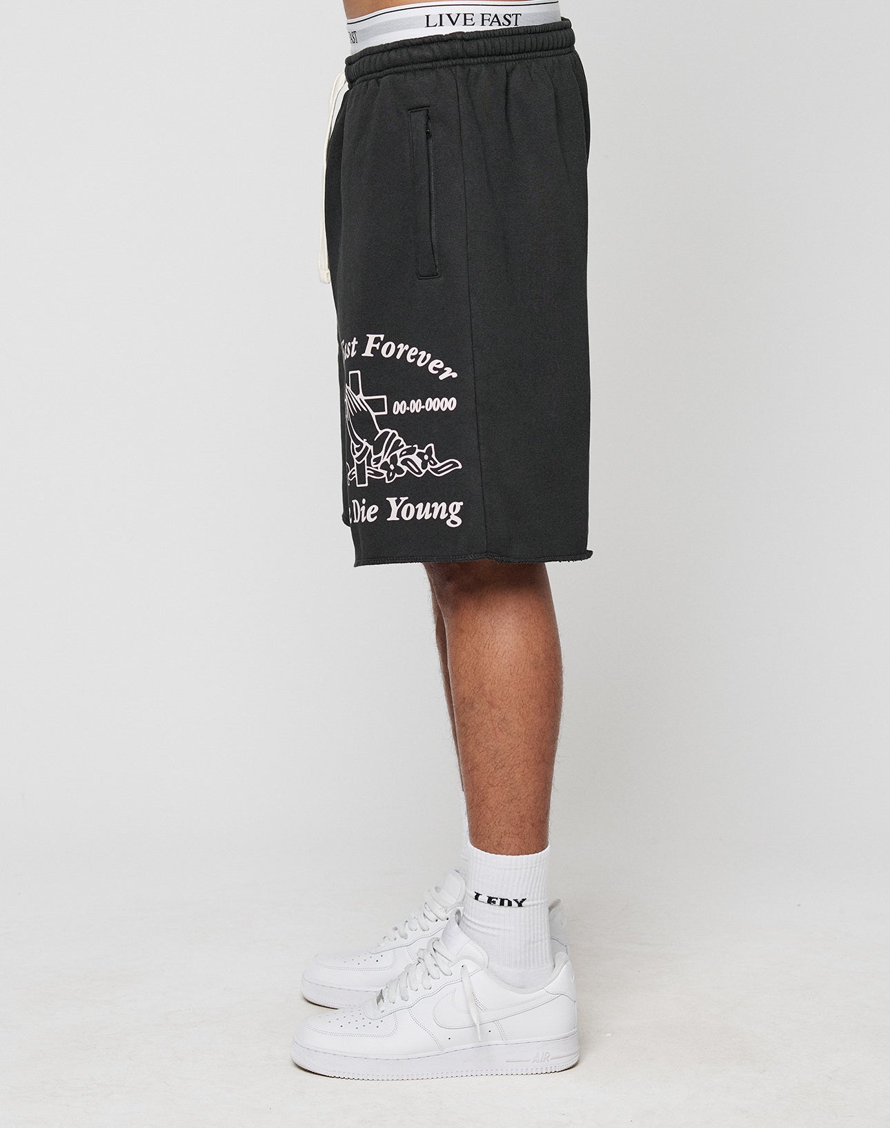 A person stands sideways wearing black LIVE FAST DIE YOUNG LFDY Praying Shorts with elastic waistband and side pockets, plus white LFDY socks and white sneakers. The shorts display a graphic with Last Forever, Die Young. Only the lower body is visible.