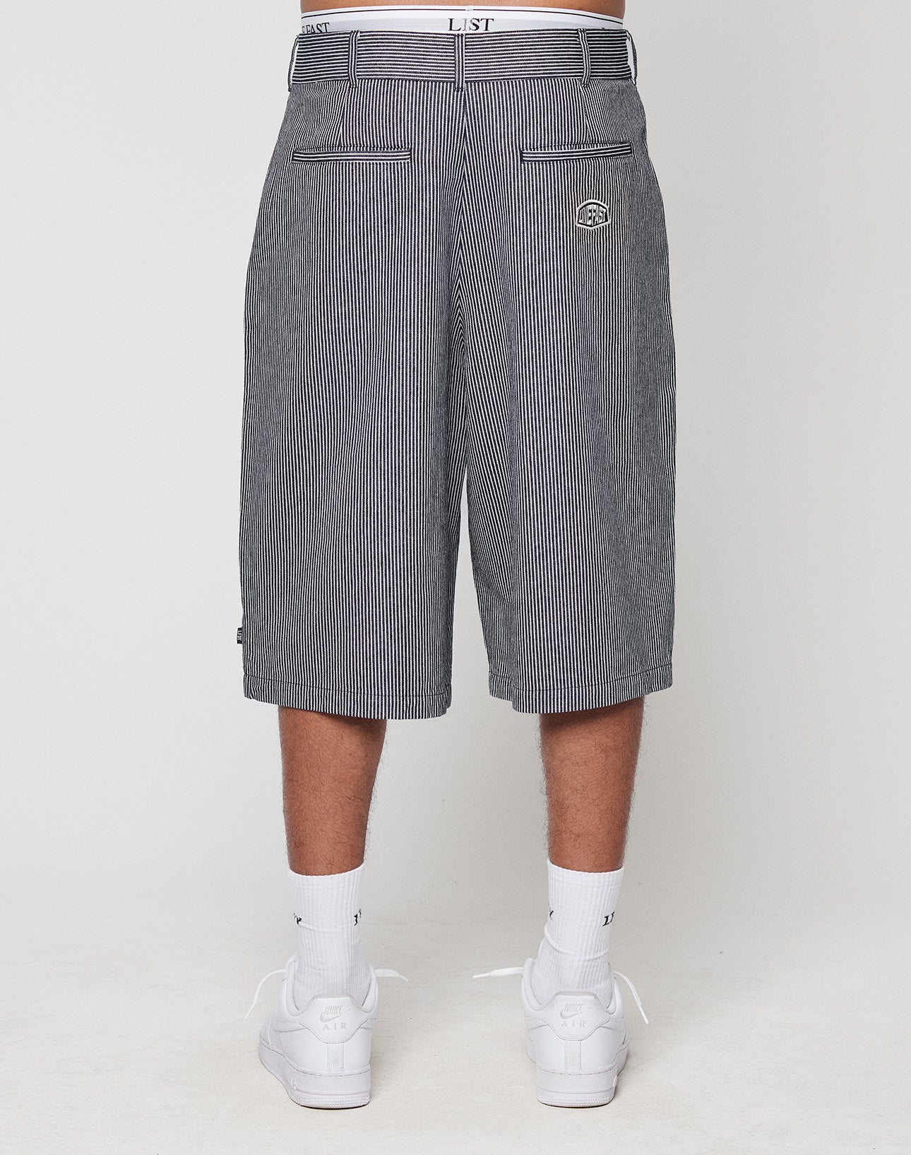 A person in pinstriped, 100% Baumwolle LFDY Twill Shorts by LIVE FAST DIE YOUNG with weites gerades Bein, two back pockets, and a small embroidered logo stands with their back to the camera, wearing white socks and sneakers.