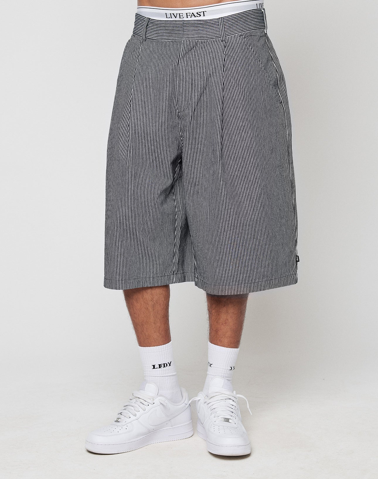 A person wears LFDY Twill Shorts by LIVE FAST DIE YOUNG with a long, black and white pinstripe design, wide straight leg, oversize fit, plus white sneakers, white LFDY socks, and a LIVE FAST waistband visible against a plain background.