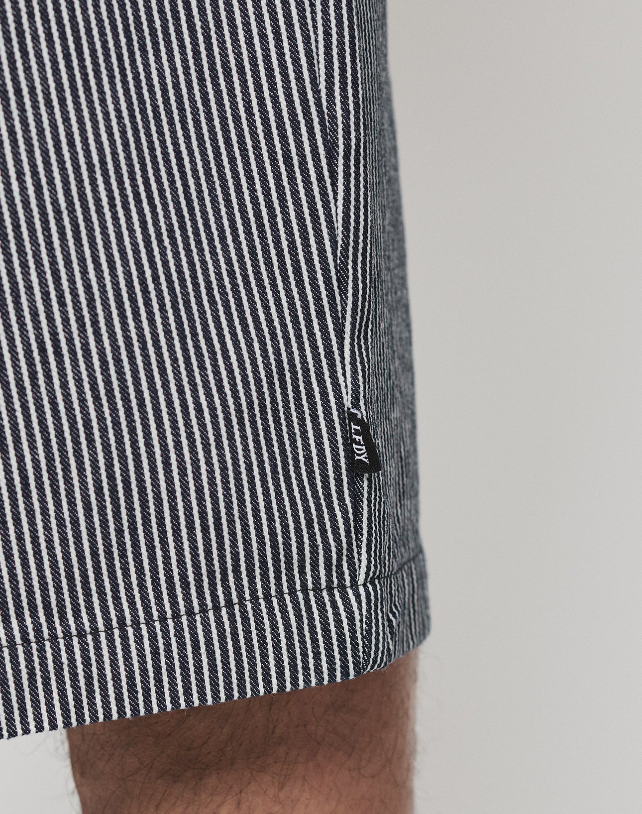 Close-up of a person wearing black and white vertically striped LFDY Twill Shorts from LIVE FAST DIE YOUNG with an oversize fit. The shorts feature a small black fabric tag on the side seam. Background is plain and light-colored.